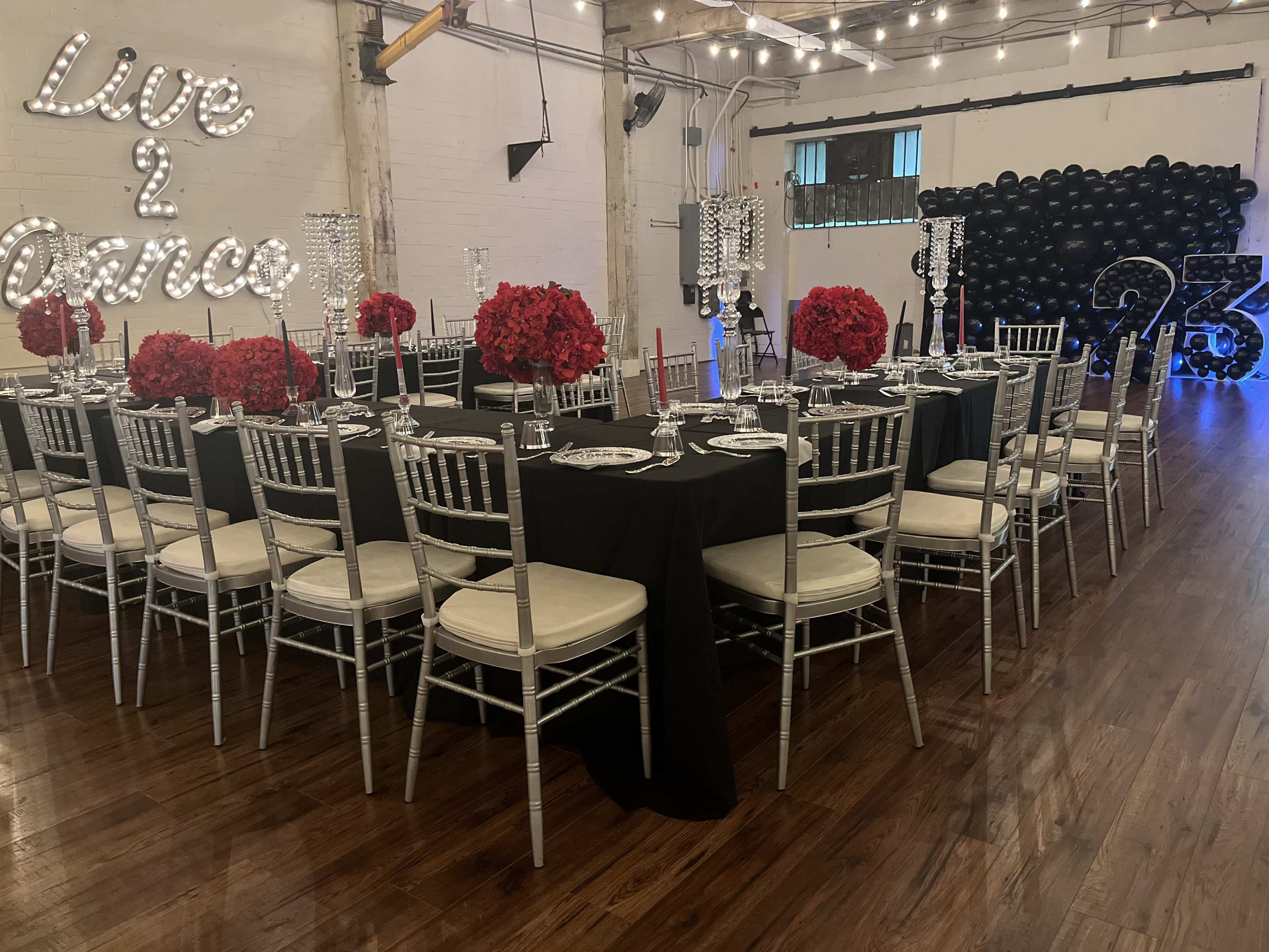 A large banquet hall is elegantly set up for an event, featuring a black tablecloth, silver chairs, red floral arrangements, and a backdrop of black balloons.
