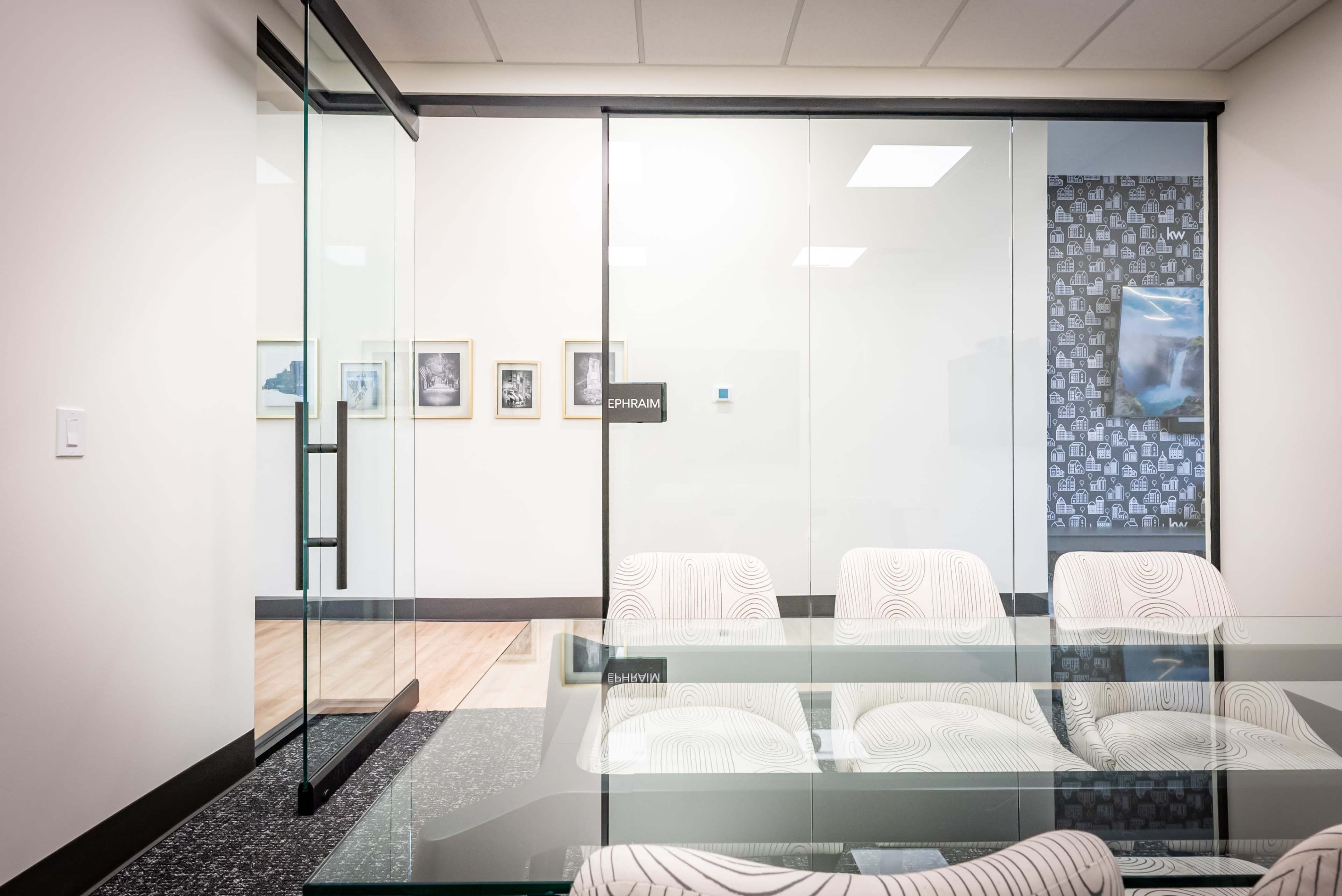 A modern conference room with a glass wall, a table, and patterned chairs, adjacent to a hallway featuring framed pictures.