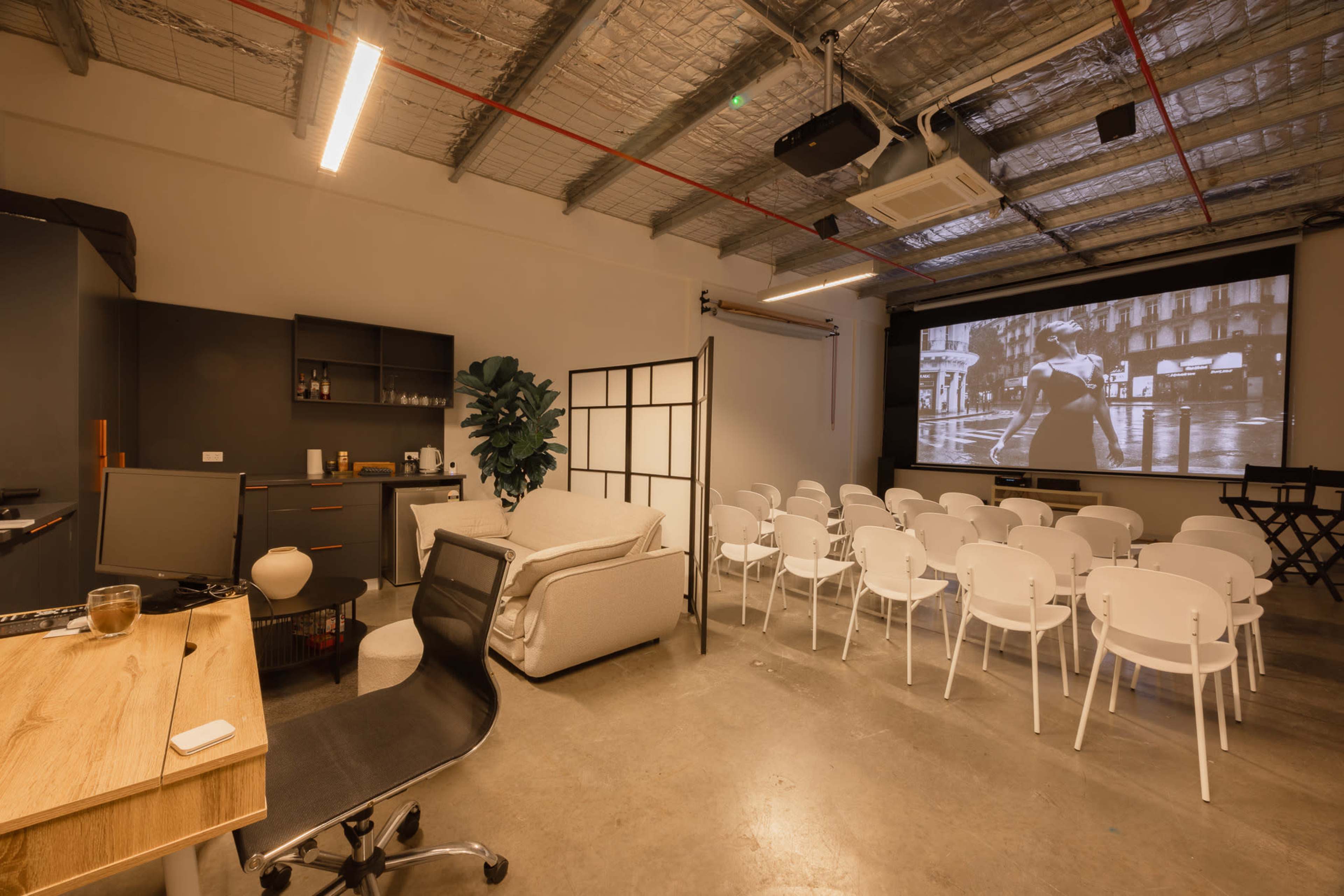 The image shows a modern meeting room with a projection screen at the front, arranged seating in white chairs, a couch, and a small kitchenette area.