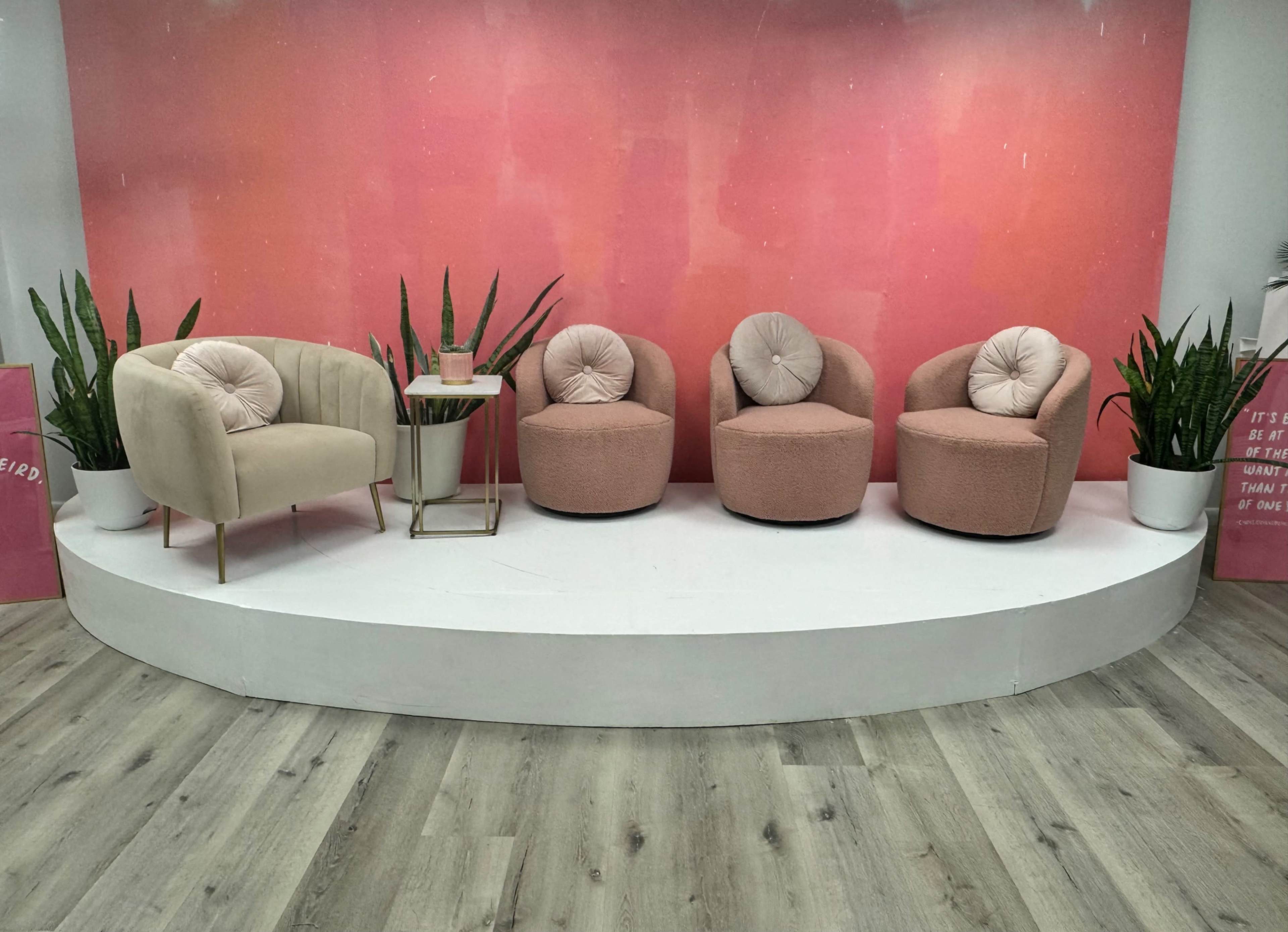 A seating arrangement featuring a beige armchair and three pink upholstered chairs, placed on a circular platform with potted plants, against a pink gradient backdrop.