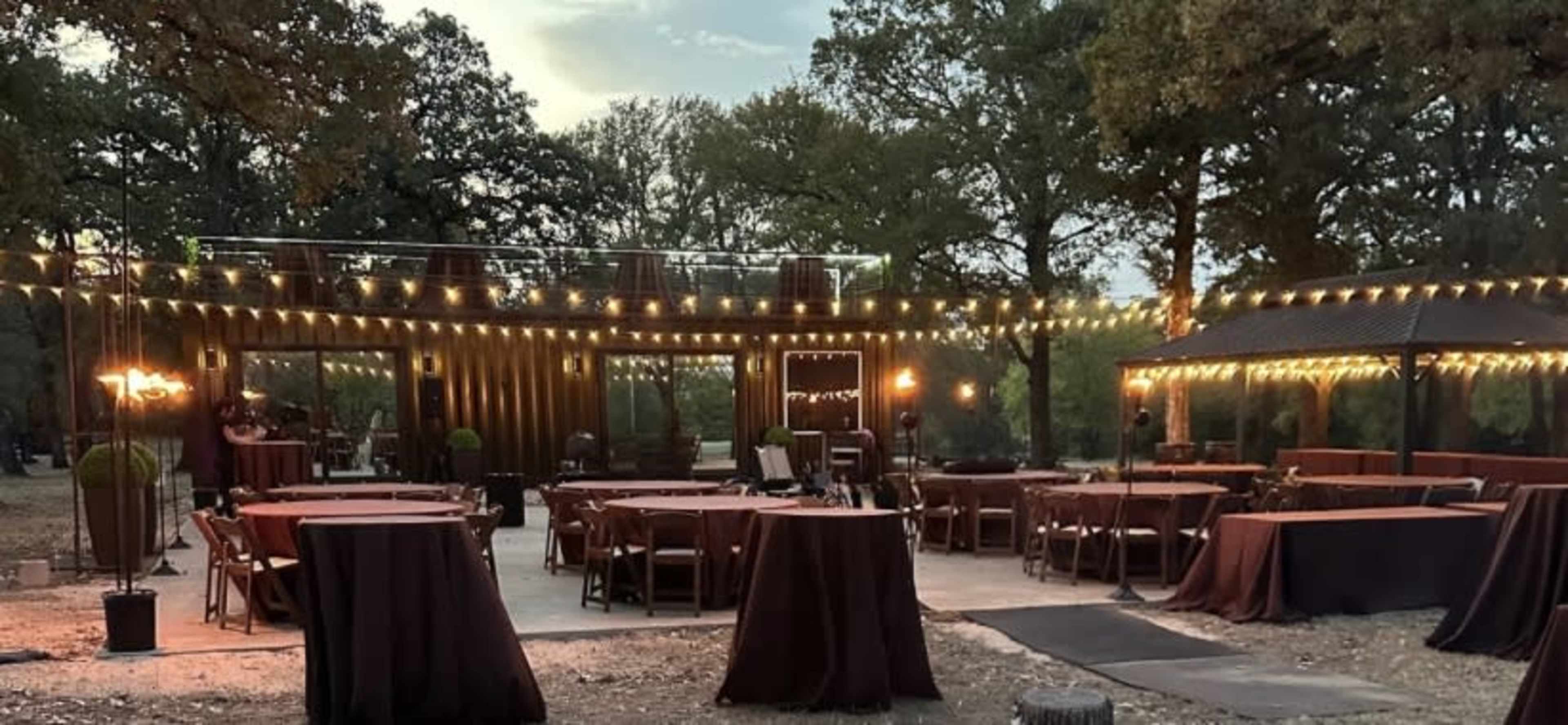 The image shows an outdoor event space with tables covered in dark cloths, string lights hanging above, and a tree-lined background.