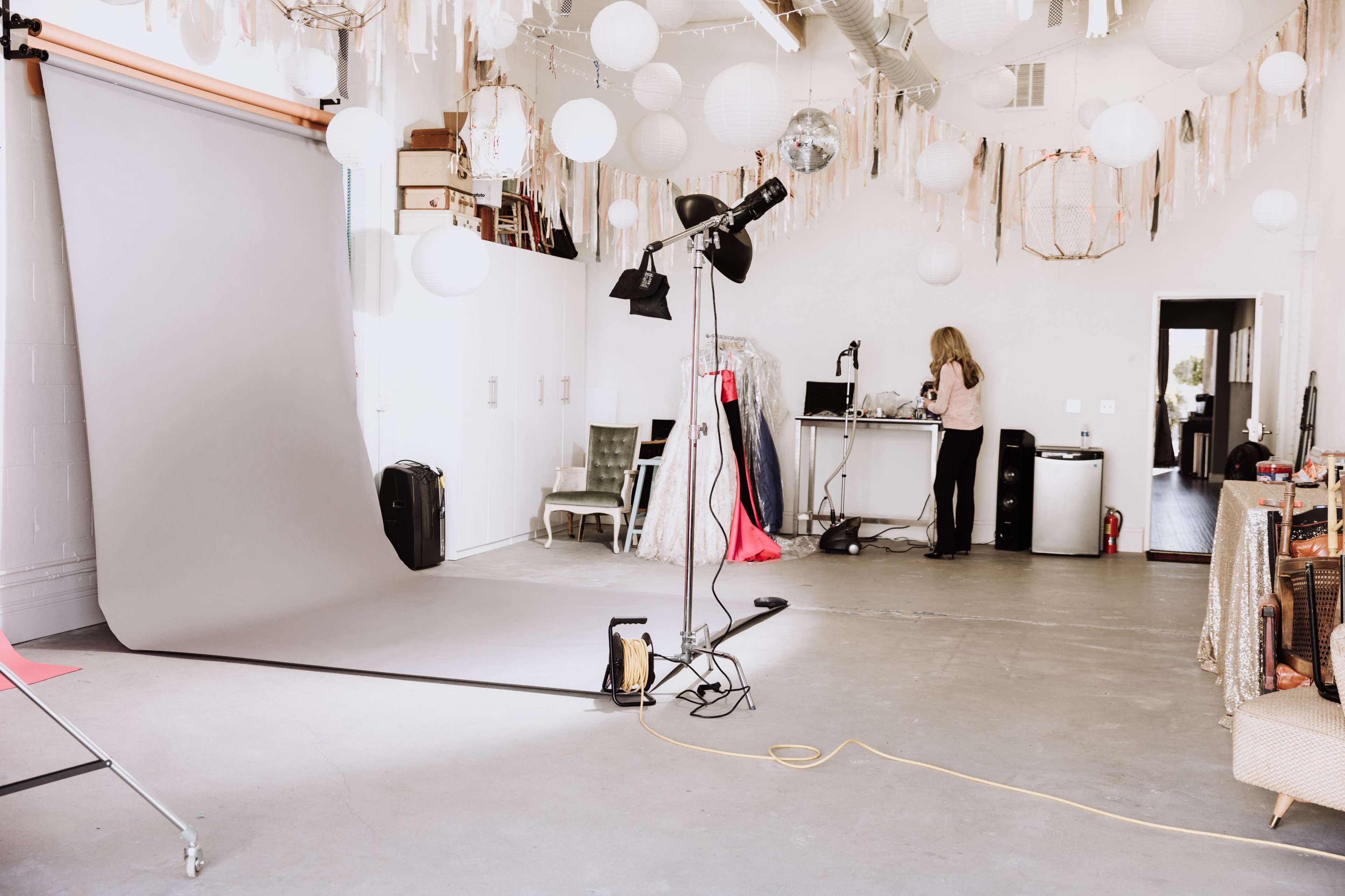 A photography studio with a gray backdrop, various decorative lanterns hanging from the ceiling, and a woman working at a desk in the background.