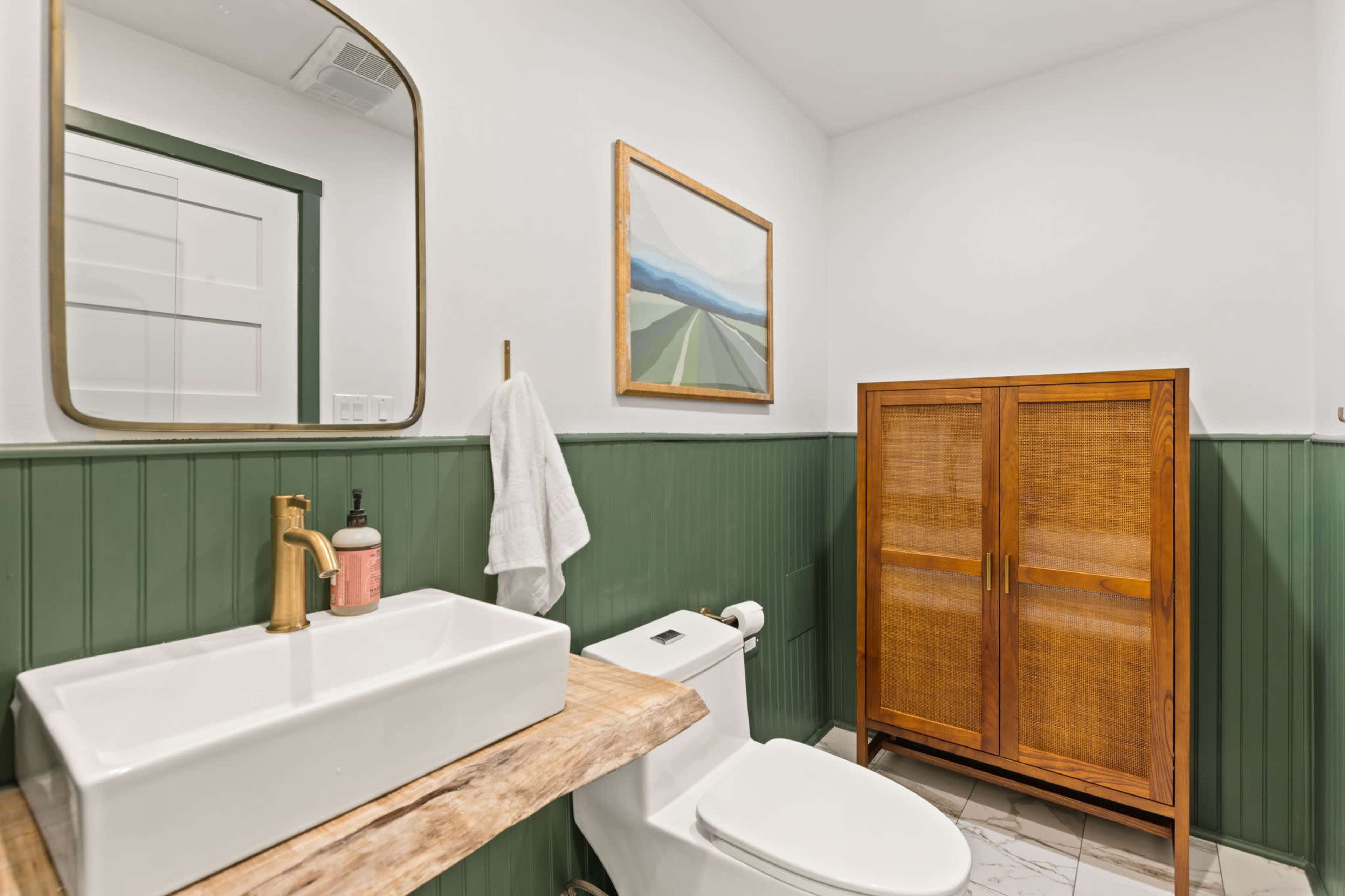 A bathroom featuring a wooden vanity with a rectangular sink, a gold faucet, a mirror, a towel hanging on a hook, a wooden cabinet, and green wainscoting.