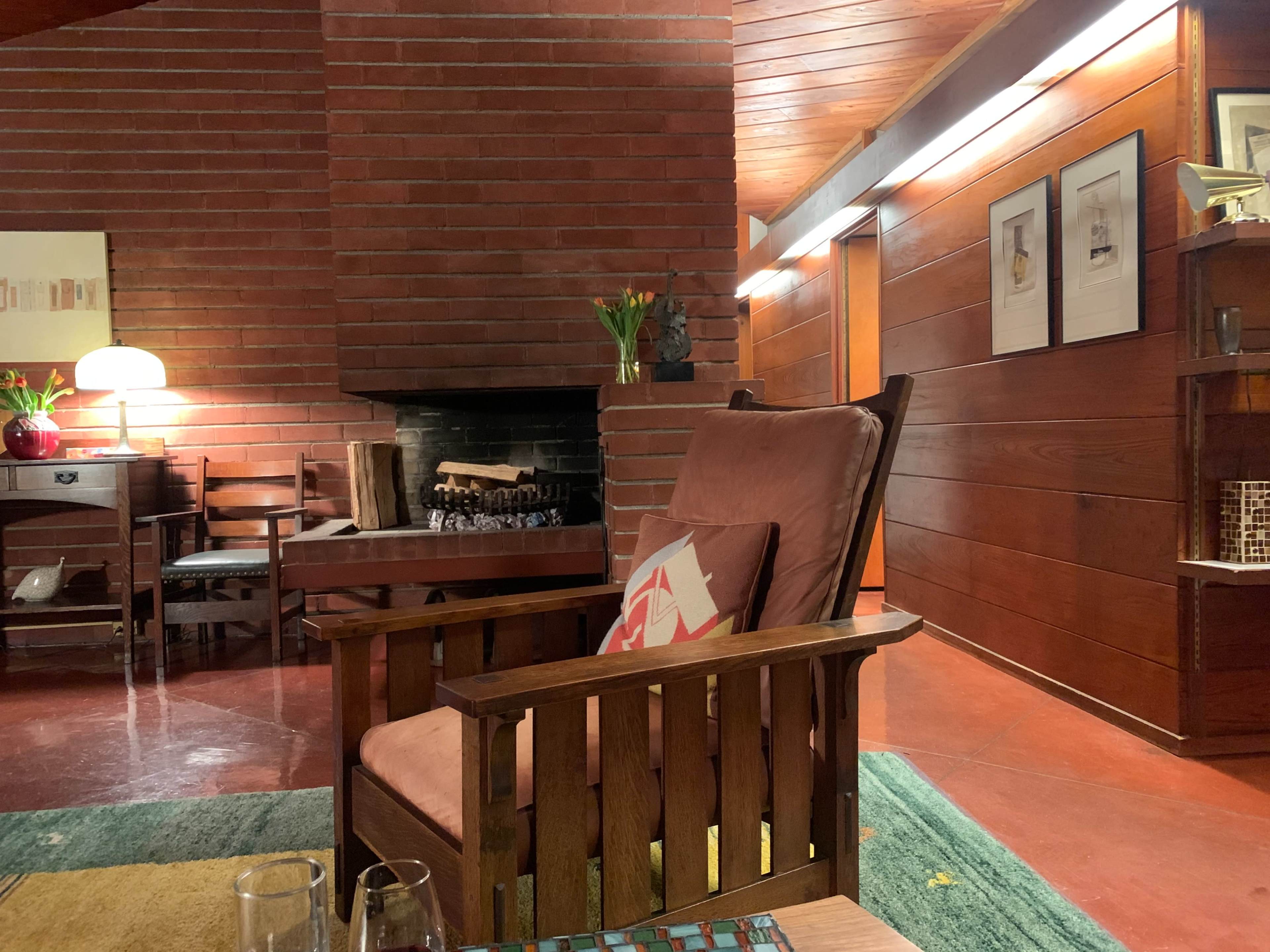 A wooden armchair with a cushion faces a brick fireplace in a room featuring wooden walls and a small table with wine glasses.