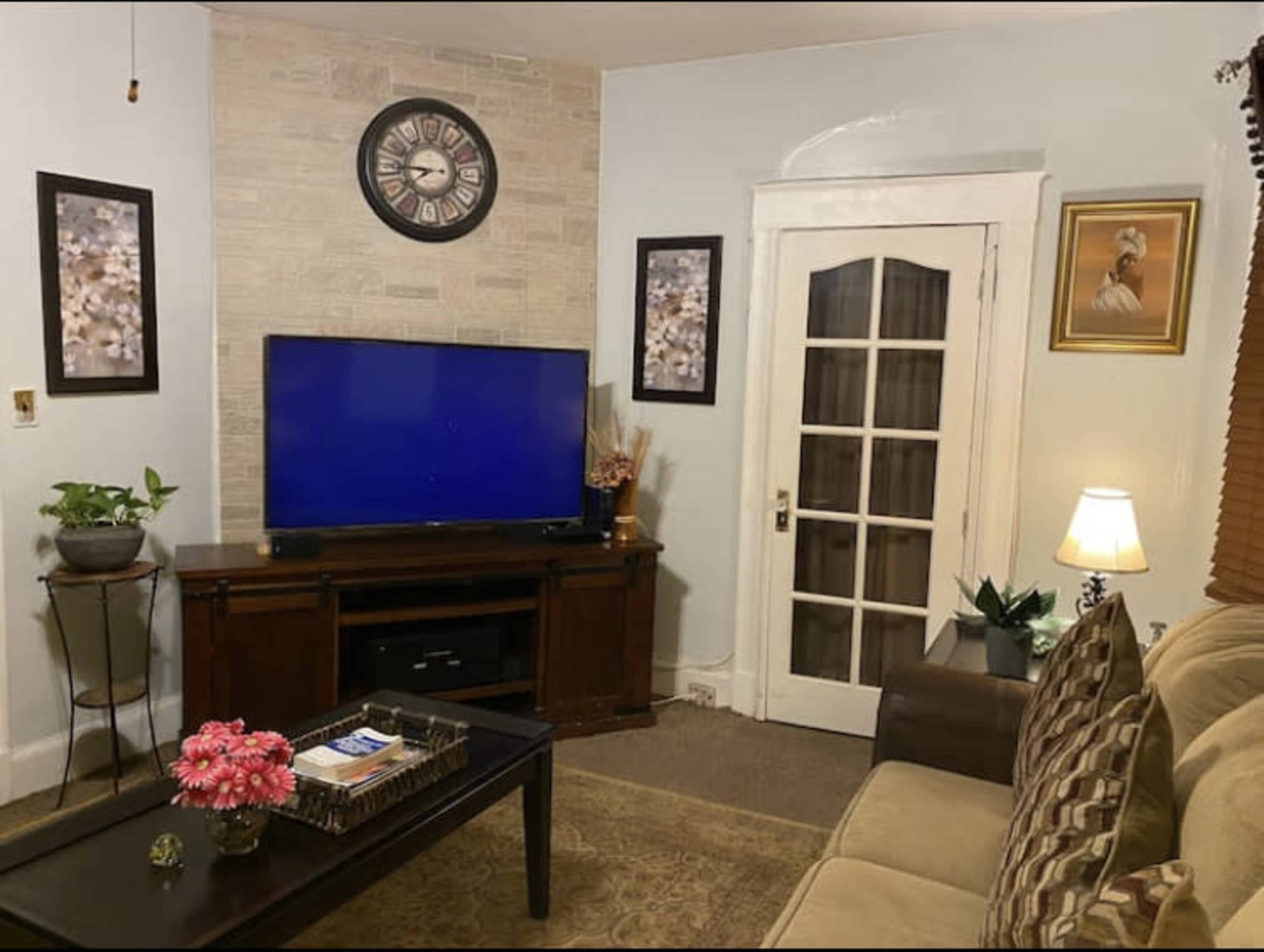 A cozy living room featuring a wall clock, a large television on a wooden stand, framed artwork, and a sofa with floral decorations on a coffee table.