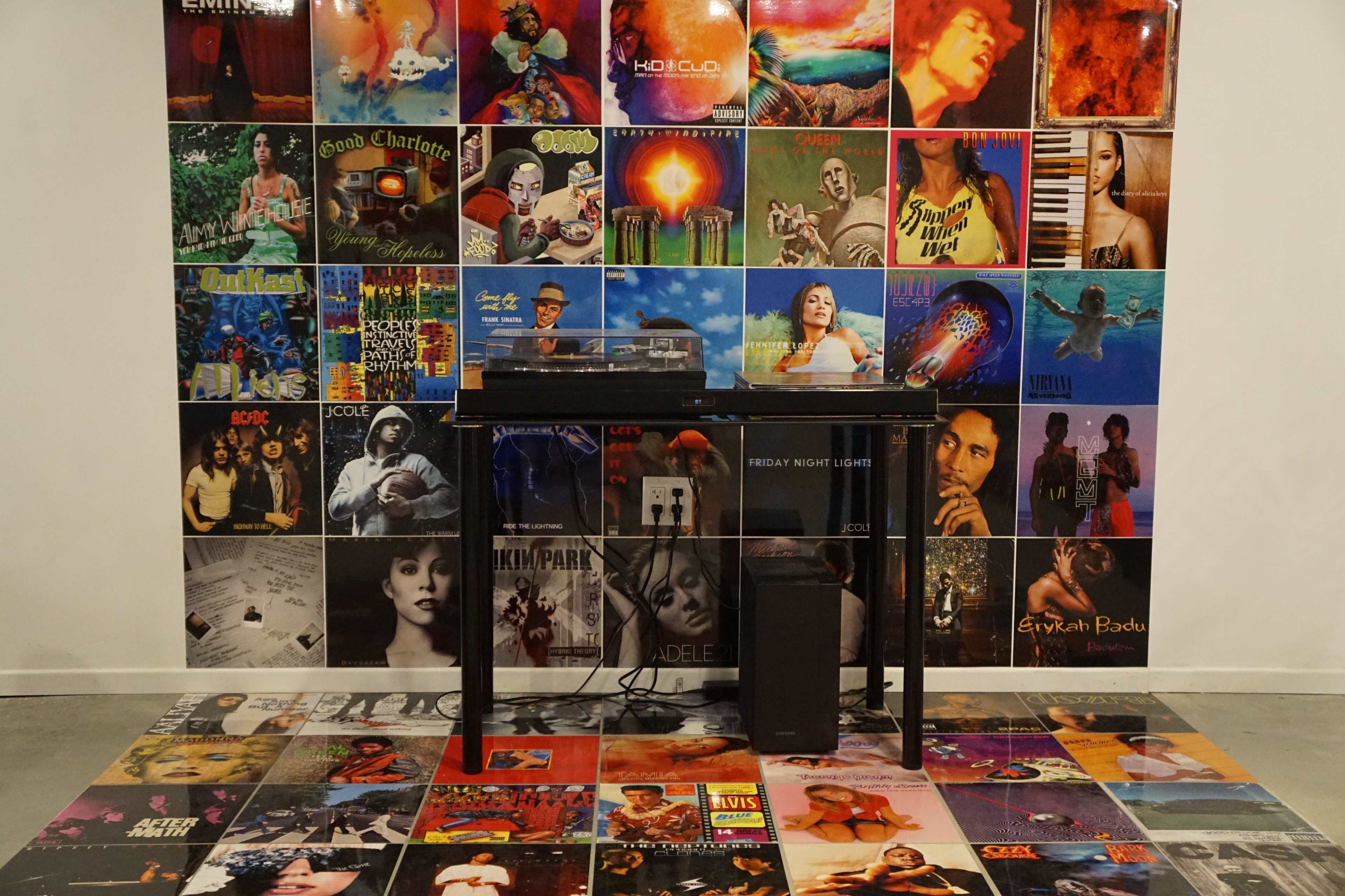A display features a wall covered in colorful vinyl record covers, with a black table holding a turntable and speakers in front of it, and more record covers laid out on the floor.