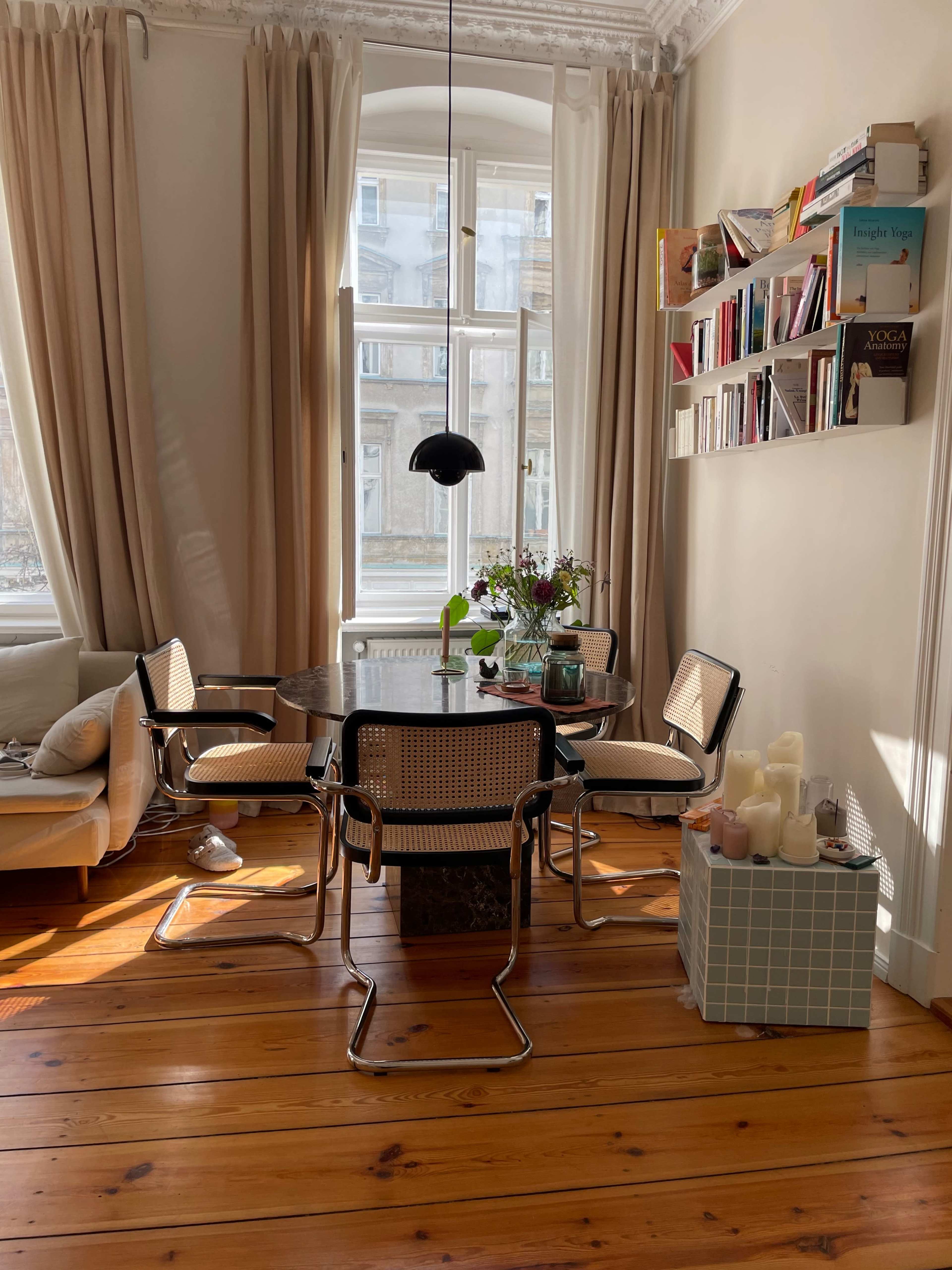 A spacious dining area with a round table surrounded by four chairs, a floor lamp, and a bookshelf filled with books, all illuminated by natural light from a large window.