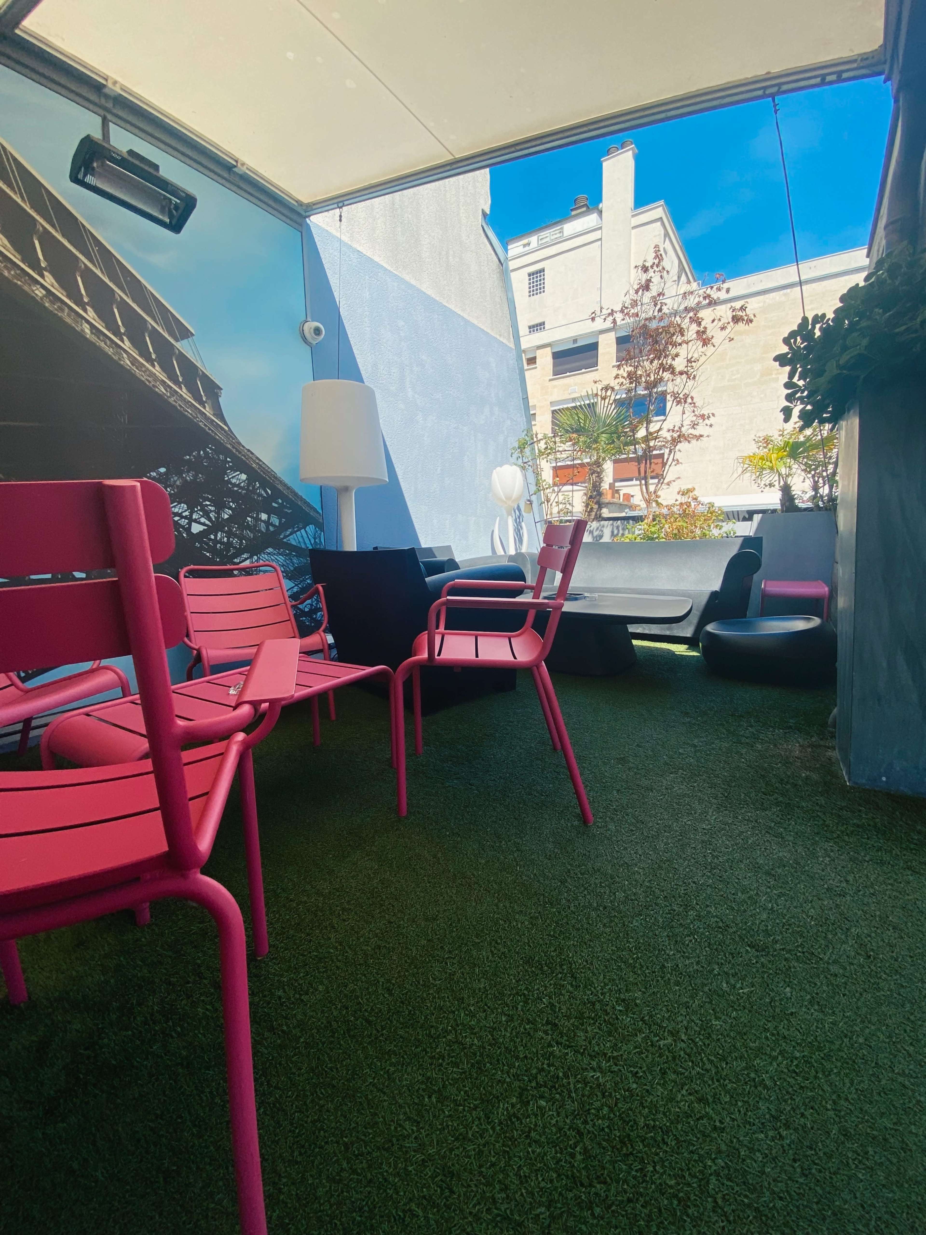 A rooftop terrace featuring pink chairs on artificial grass, with buildings visible in the background under a clear blue sky.