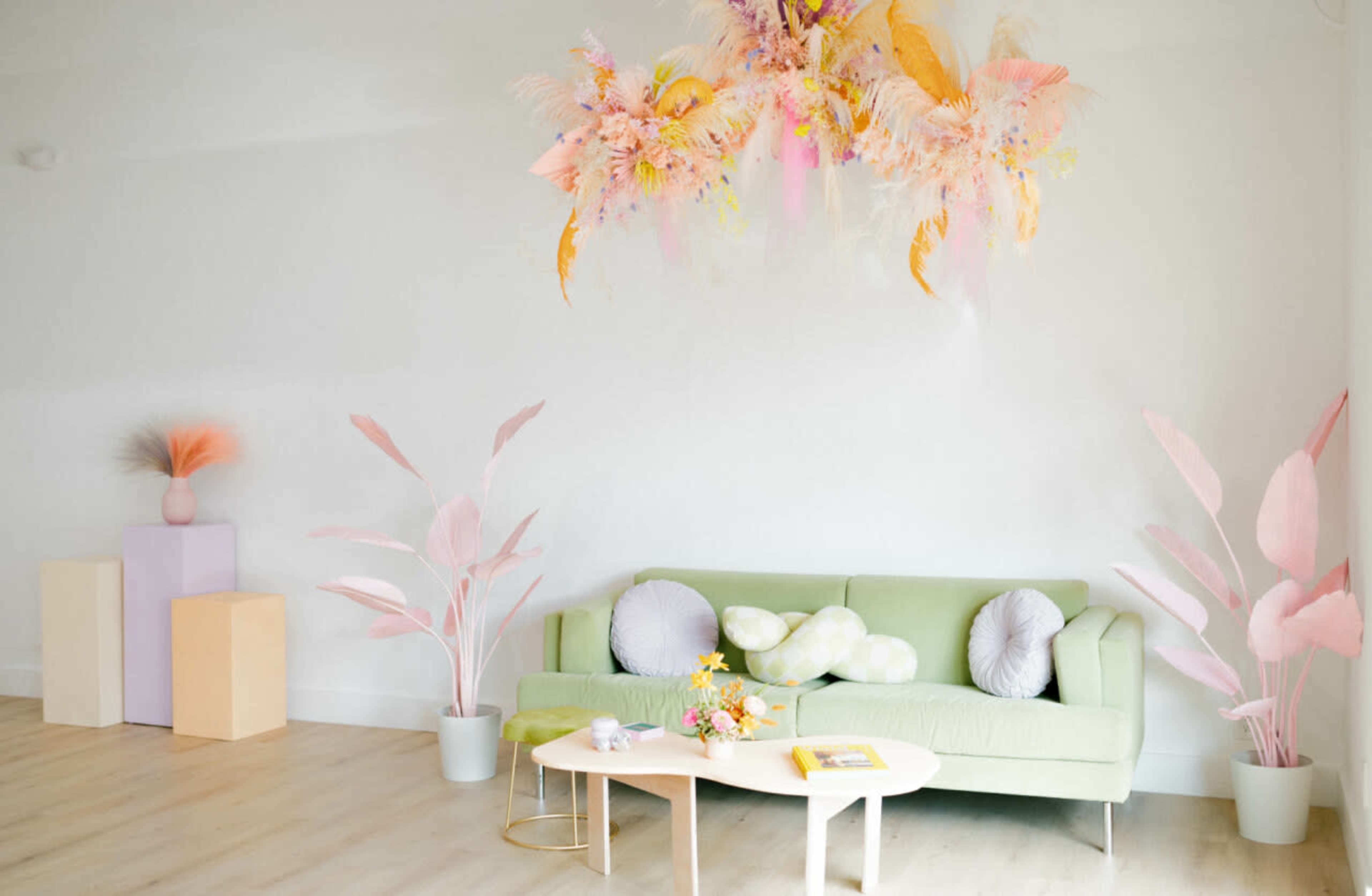 A light-filled room features a green sofa with decorative pillows, a coffee table with flowers, and pastel-colored plants and decor accents.
