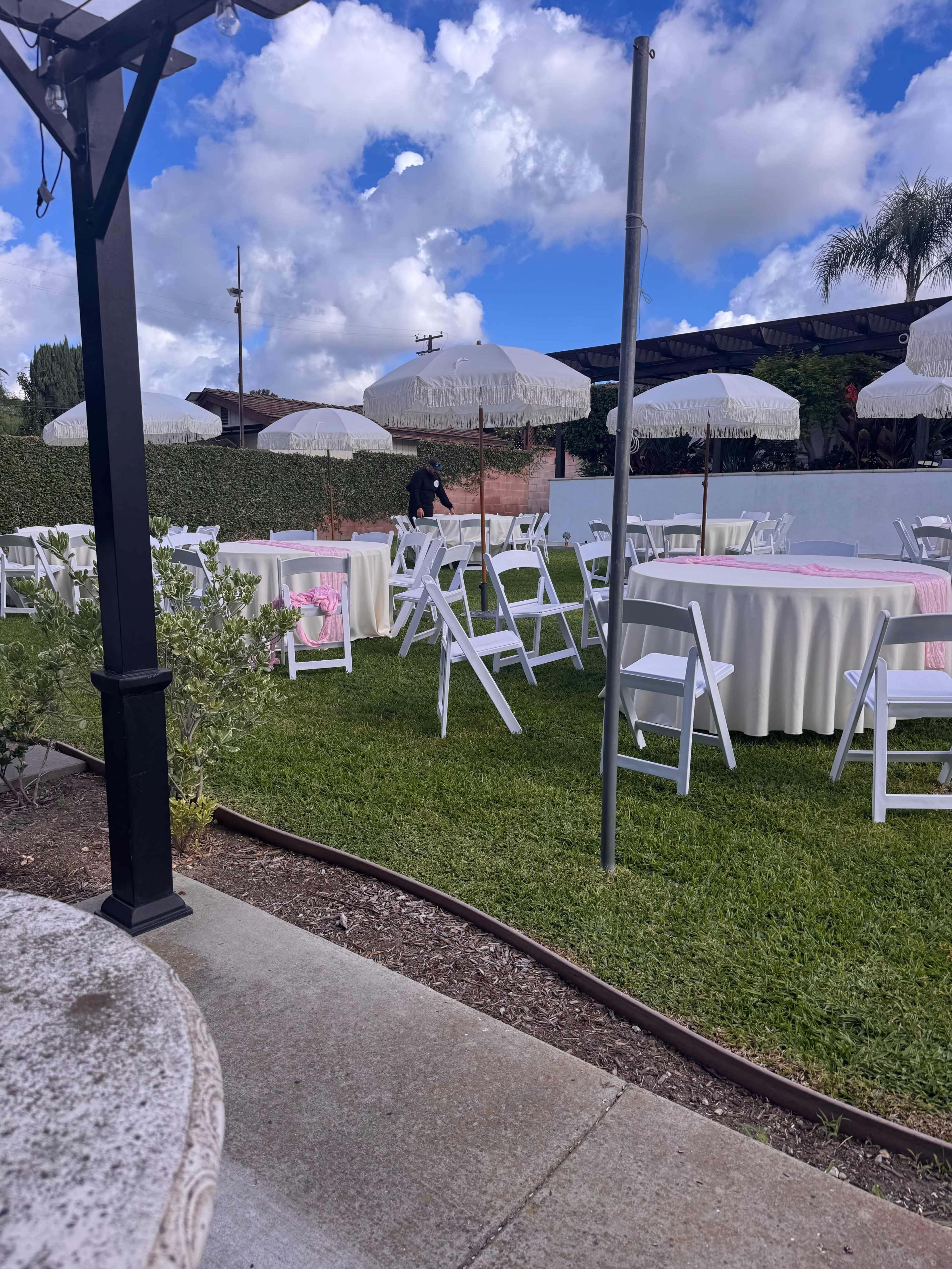 The image shows a garden event setup with white chairs and tables draped in light-colored linens, under large umbrellas, amid lush green grass and a partly cloudy sky.