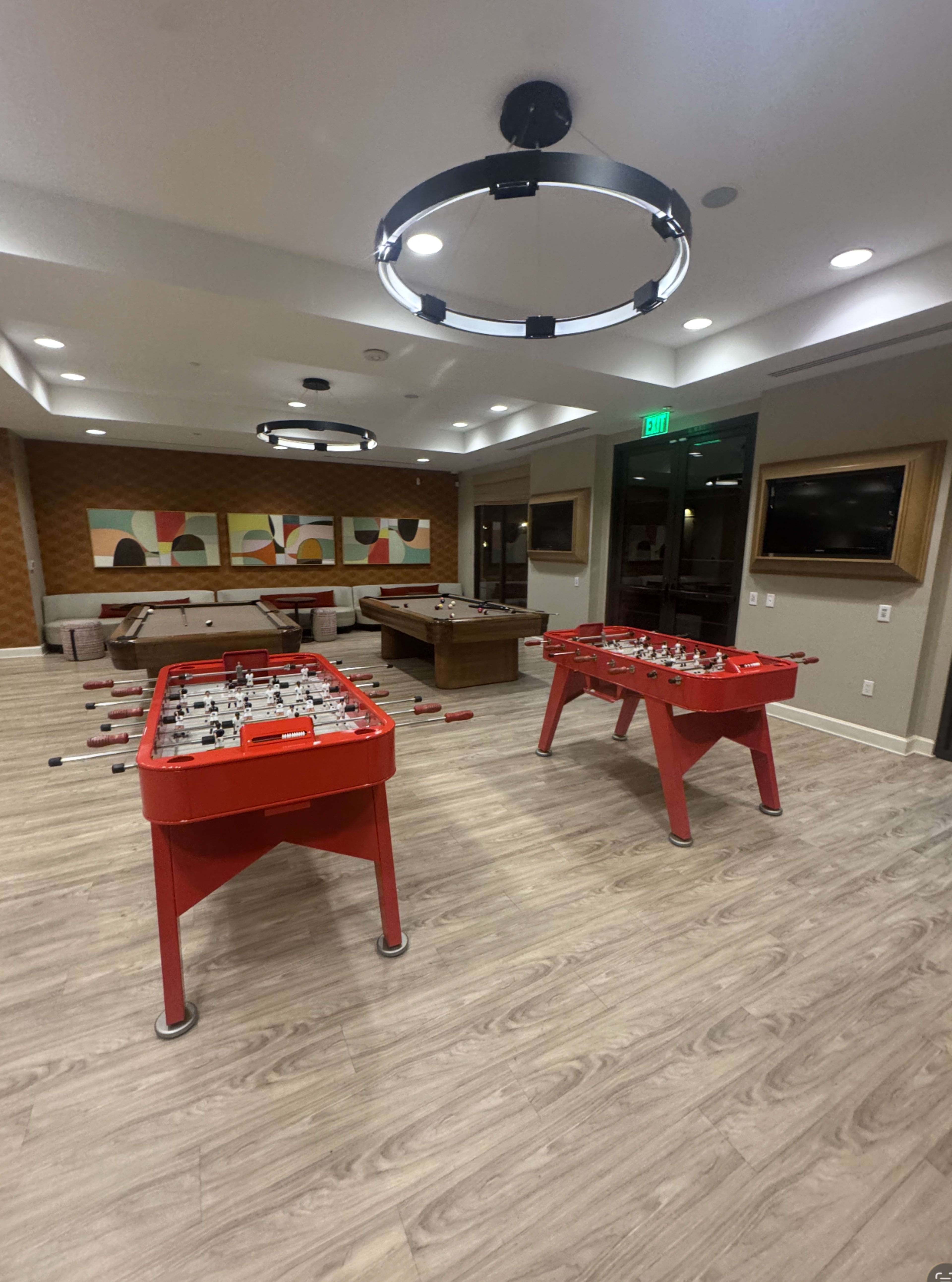 The image shows a recreation room with two red foosball tables and a pool table, featuring modern lighting and colorful artwork on the walls.