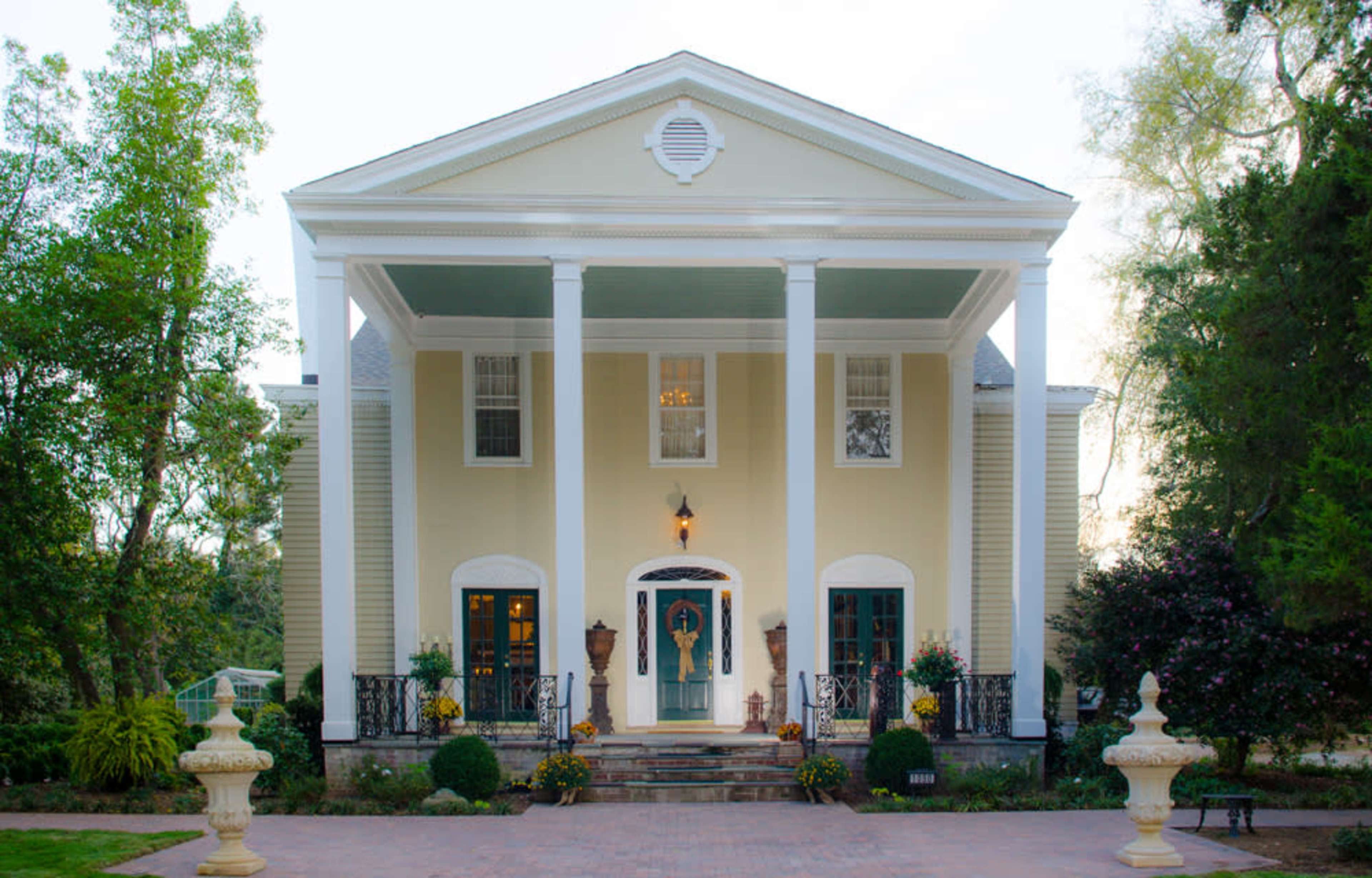 A large, two-story house with a prominent pediment and four white columns at the entrance is surrounded by landscaped gardens.