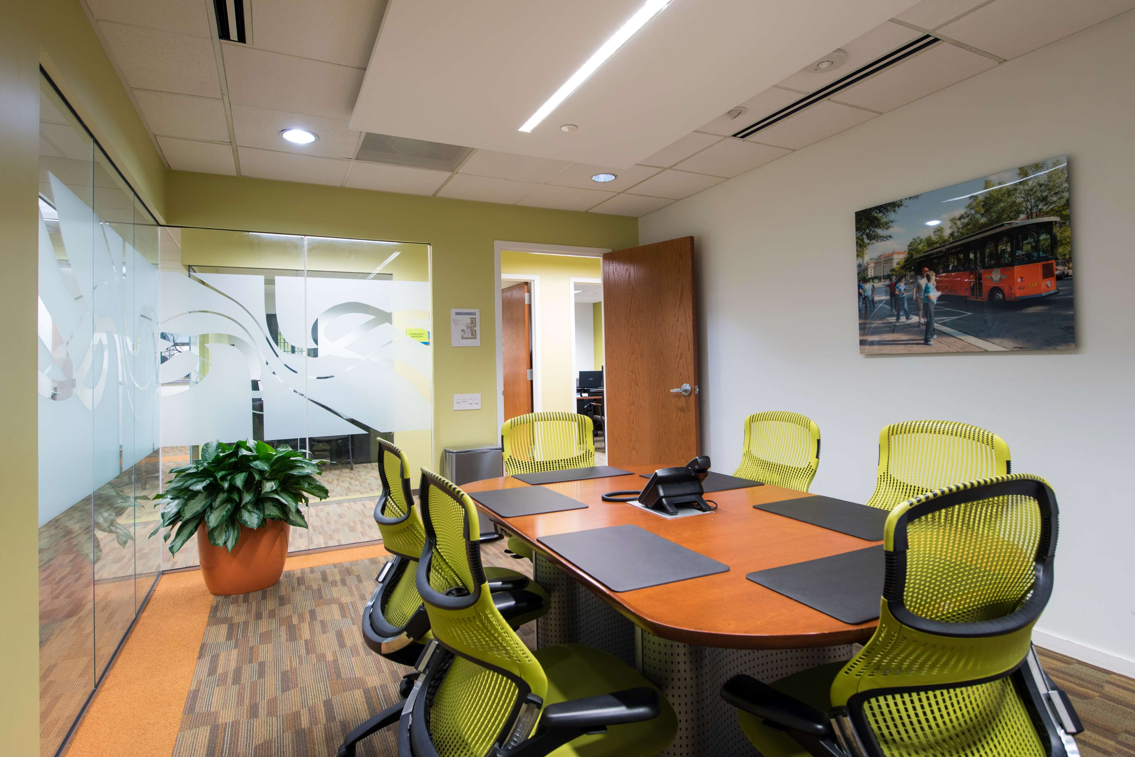 A modern conference room equipped with a wooden table, ergonomic chairs, a phone, and a large glass wall.