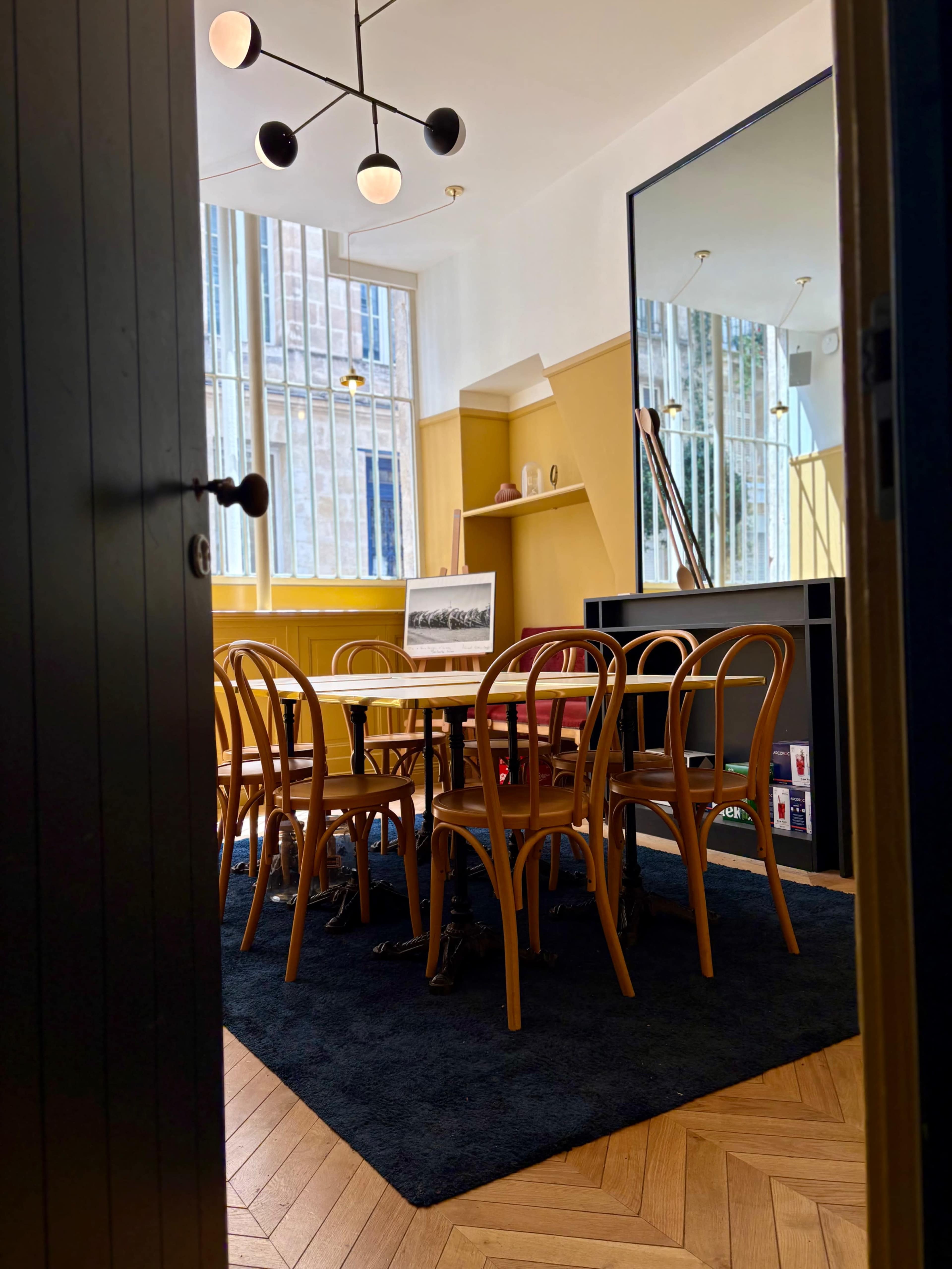 A brightly lit room features a large table surrounded by wooden chairs, with a large mirror and shelves against the walls.