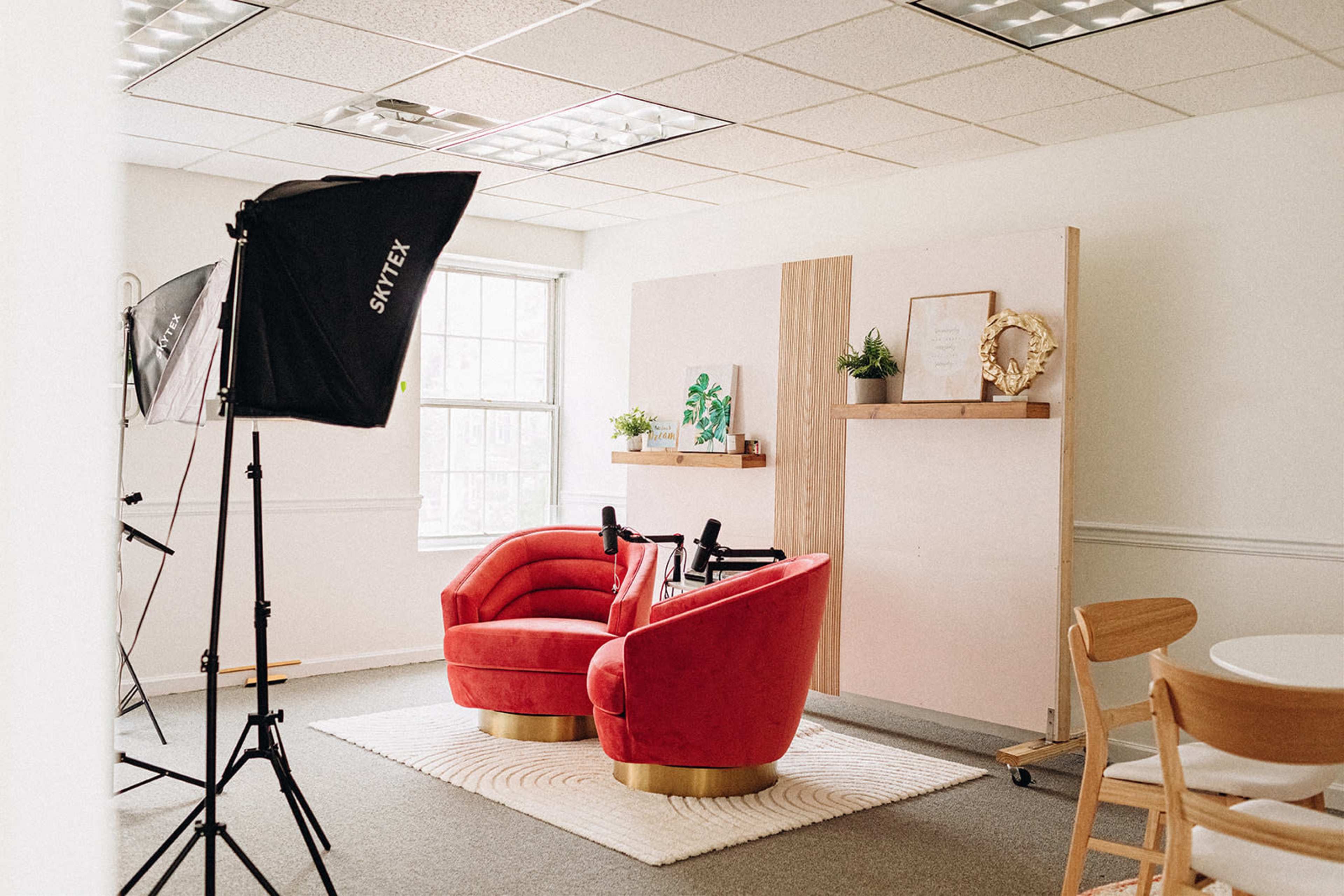 A bright room with two red velvet chairs on a circular rug, framed by a photography light setup and decorative plants on shelves.