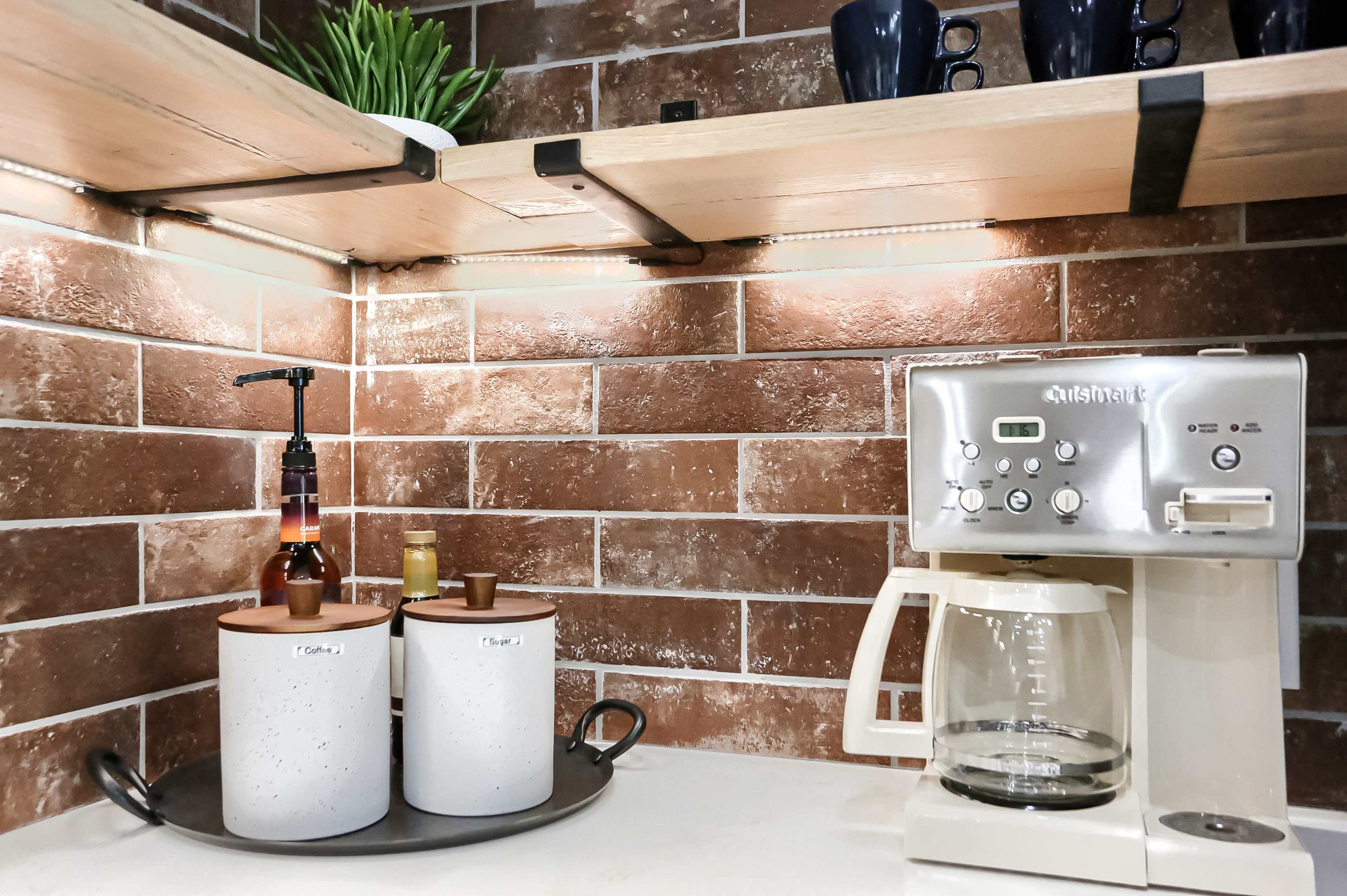 The image shows a kitchen corner with a coffee maker next to round containers on a tray, all set against a brown brick wall and illuminated by shelf lights.