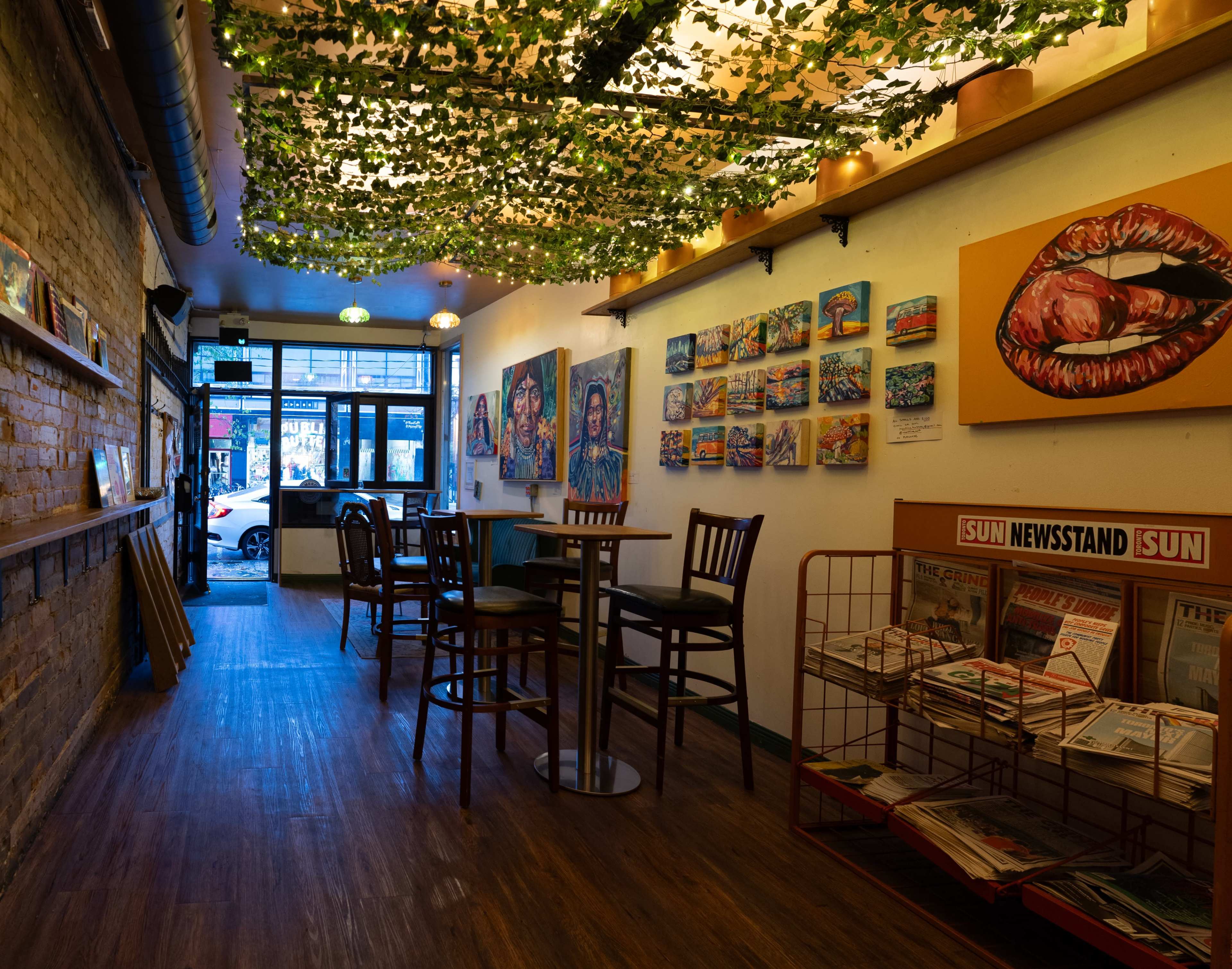 The interior of a cafe features wooden tables and chairs, vibrant artwork on the walls, and a display of newspapers near the entrance.
