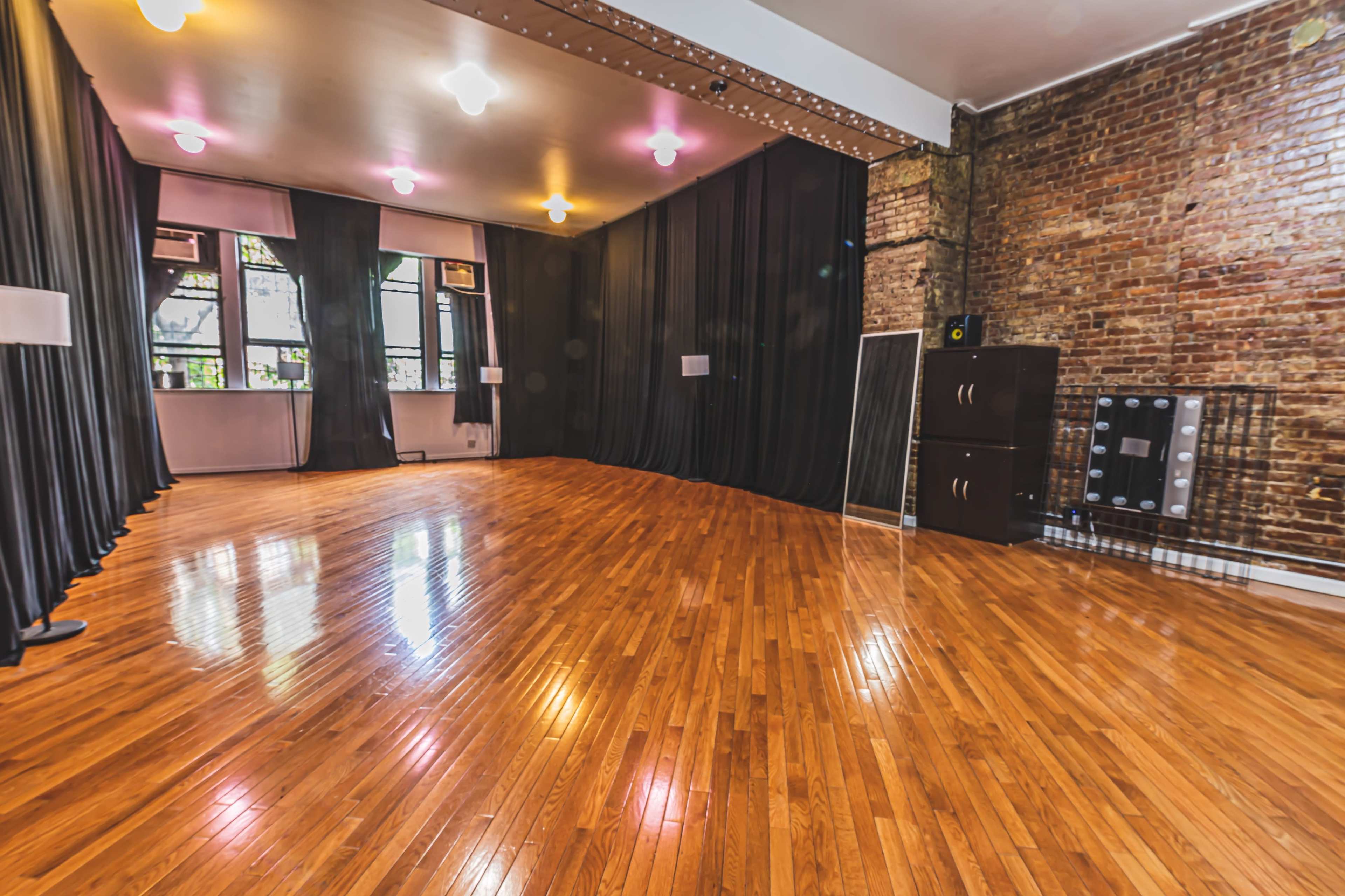 The image shows a spacious, empty room with polished hardwood flooring, exposed brick walls, and large windows draped with dark curtains.