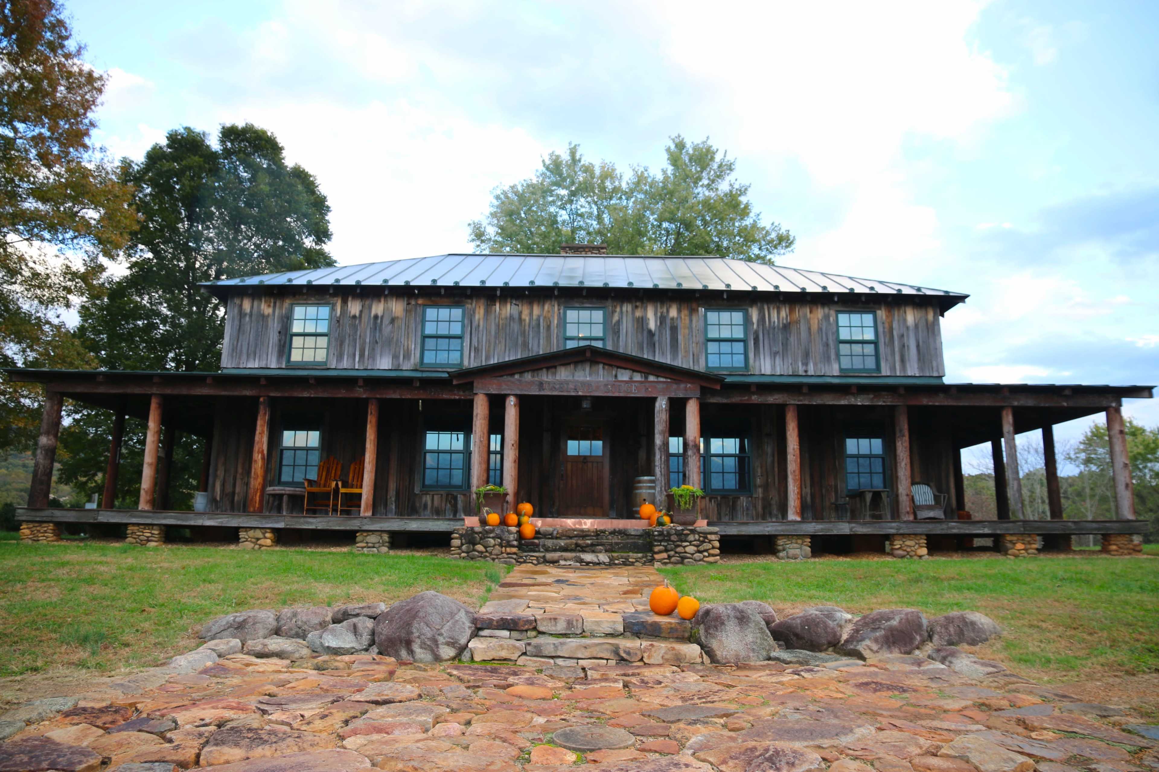 A rustic two-story wooden house with a metal roof is surrounded by rocks and pumpkins in the front yard.