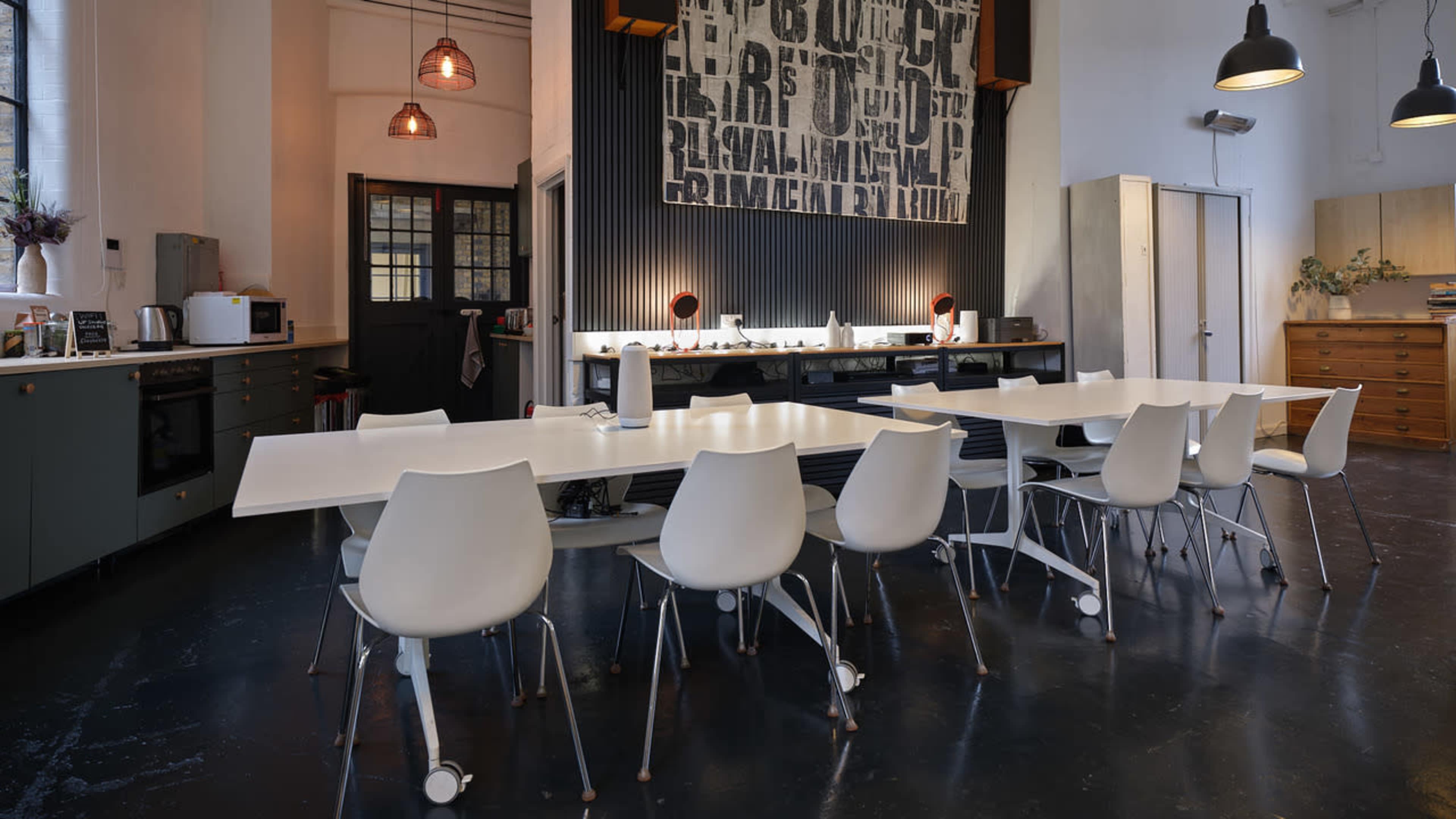 The image shows a modern kitchen and dining area featuring a long white table surrounded by white chairs, with pendant lights hanging overhead and a large wall art piece in the background.