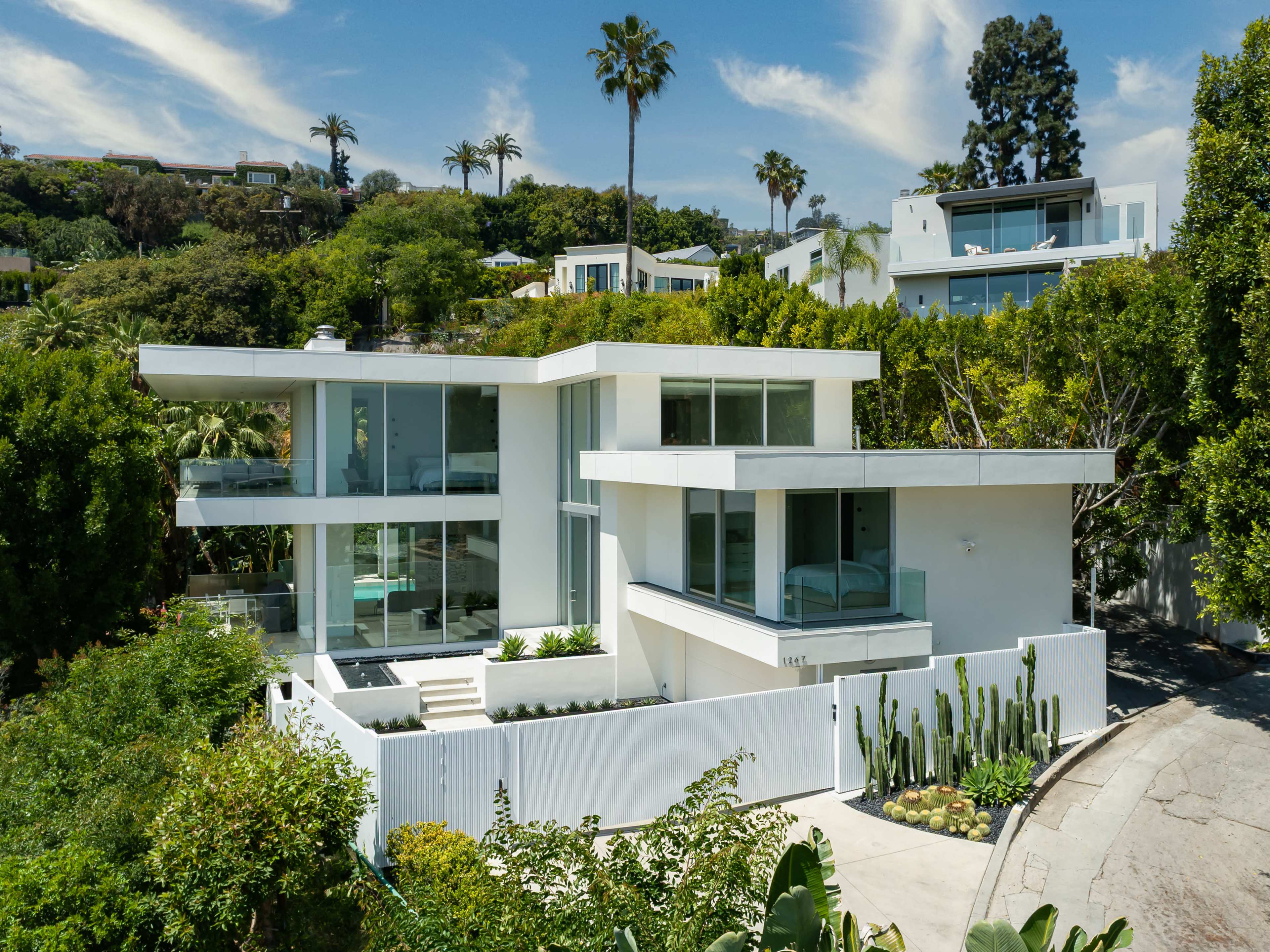 The image shows a modern two-story white house with large glass windows, surrounded by lush greenery and palm trees on a hillside.