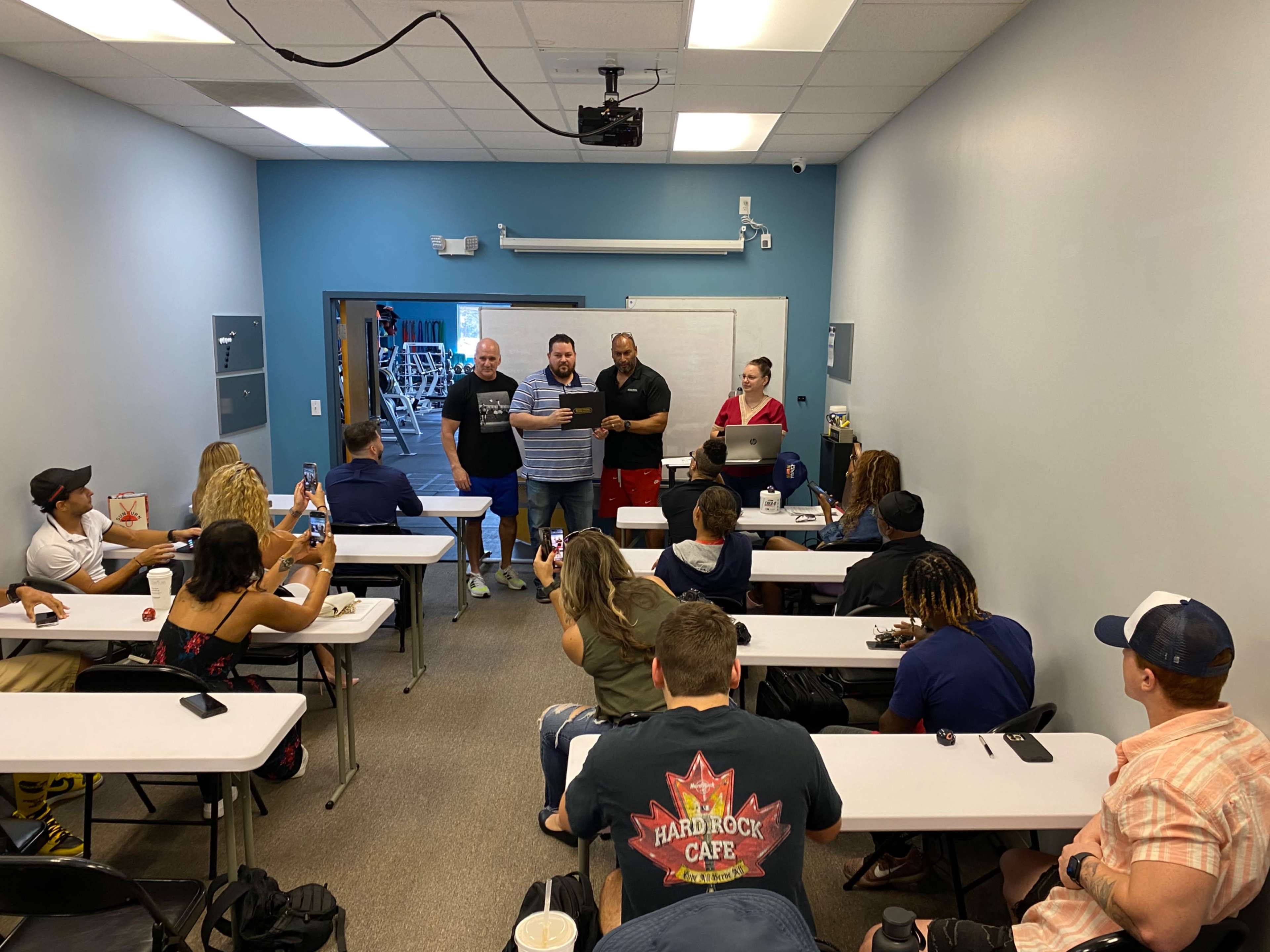 A group of adults is gathered in a classroom setting, with several people seated at tables and a few standing at the front presenting a certificate or award.