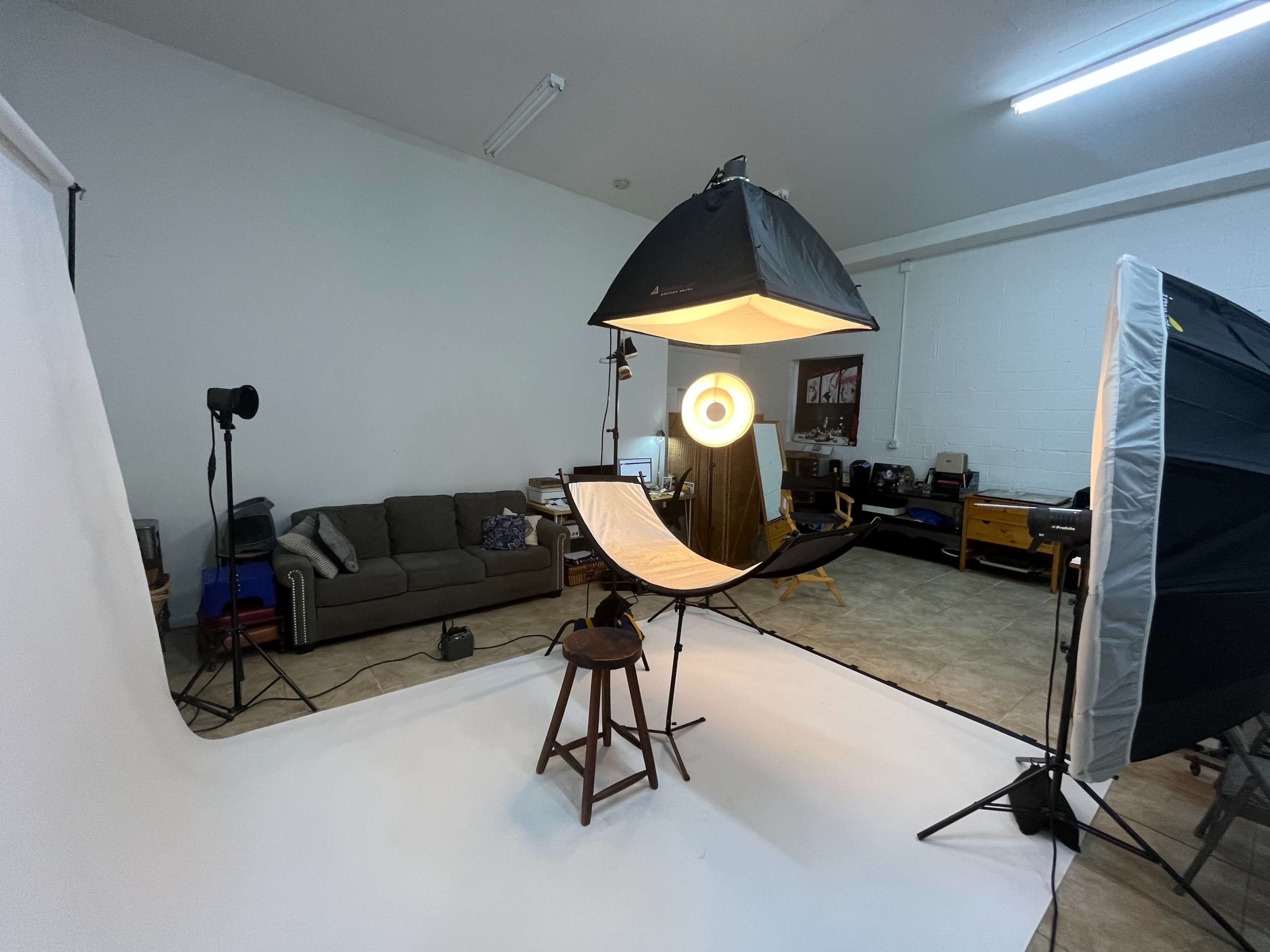 A photography setup with a softbox light, a backdrop, and a stool in a studio space that includes a couch and various equipment.