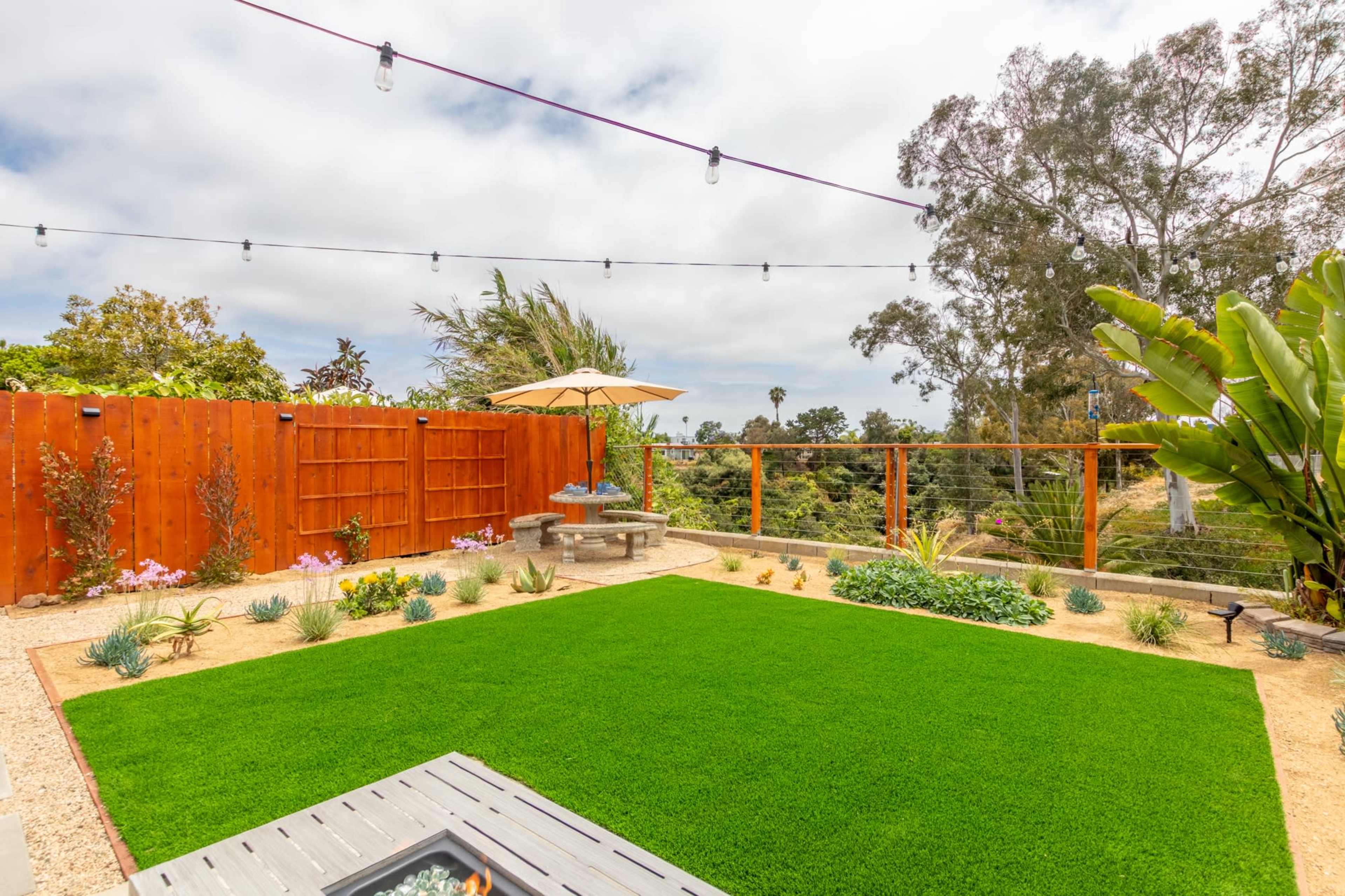 A landscaped backyard with a well-maintained lawn, surrounded by a wooden fence and a glass railing, featuring a table under an umbrella and decorative plants.