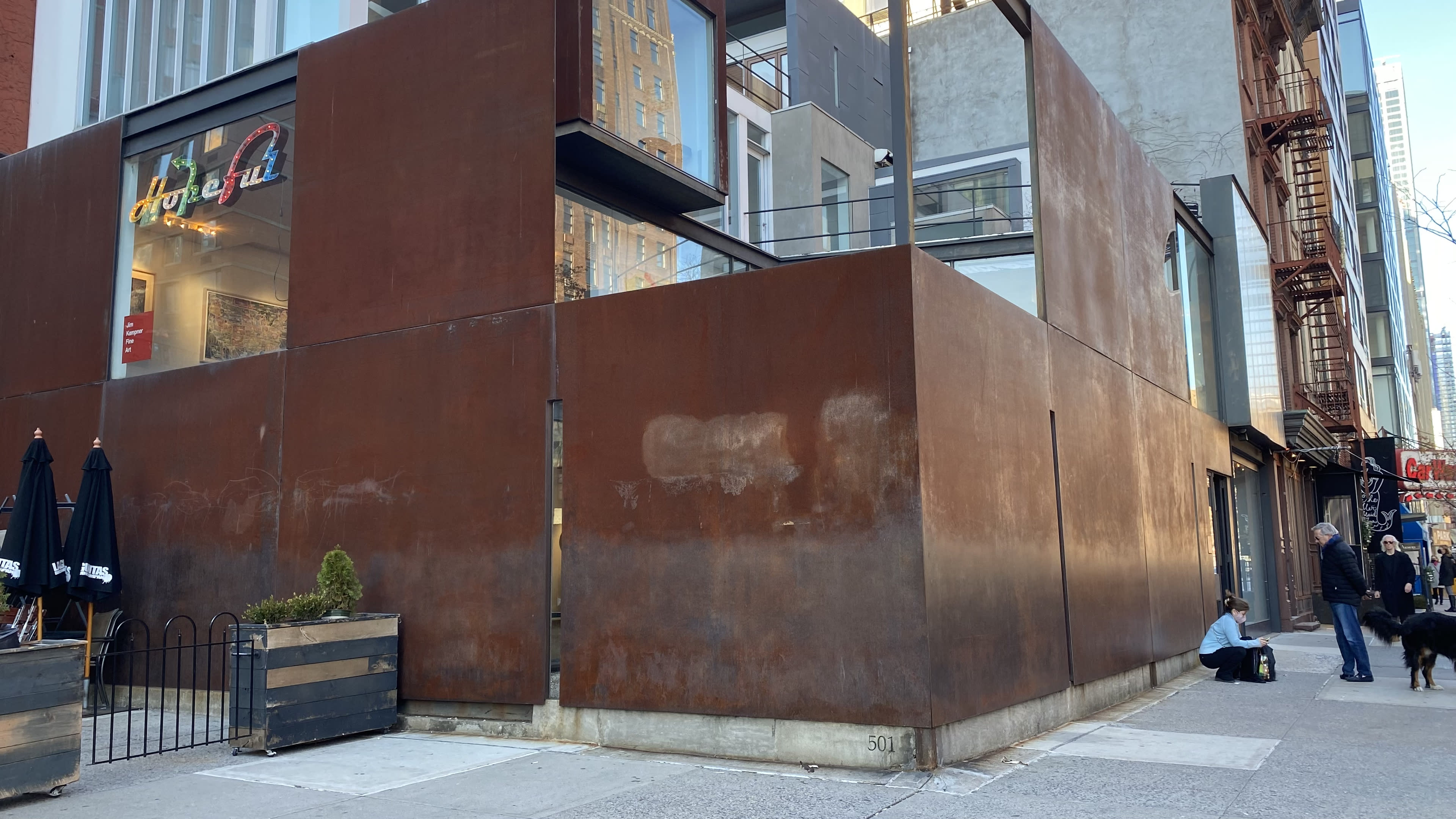 A modern building with a rust-colored metal facade stands at the corner of a city street, with large windows and nearby sidewalk seating.