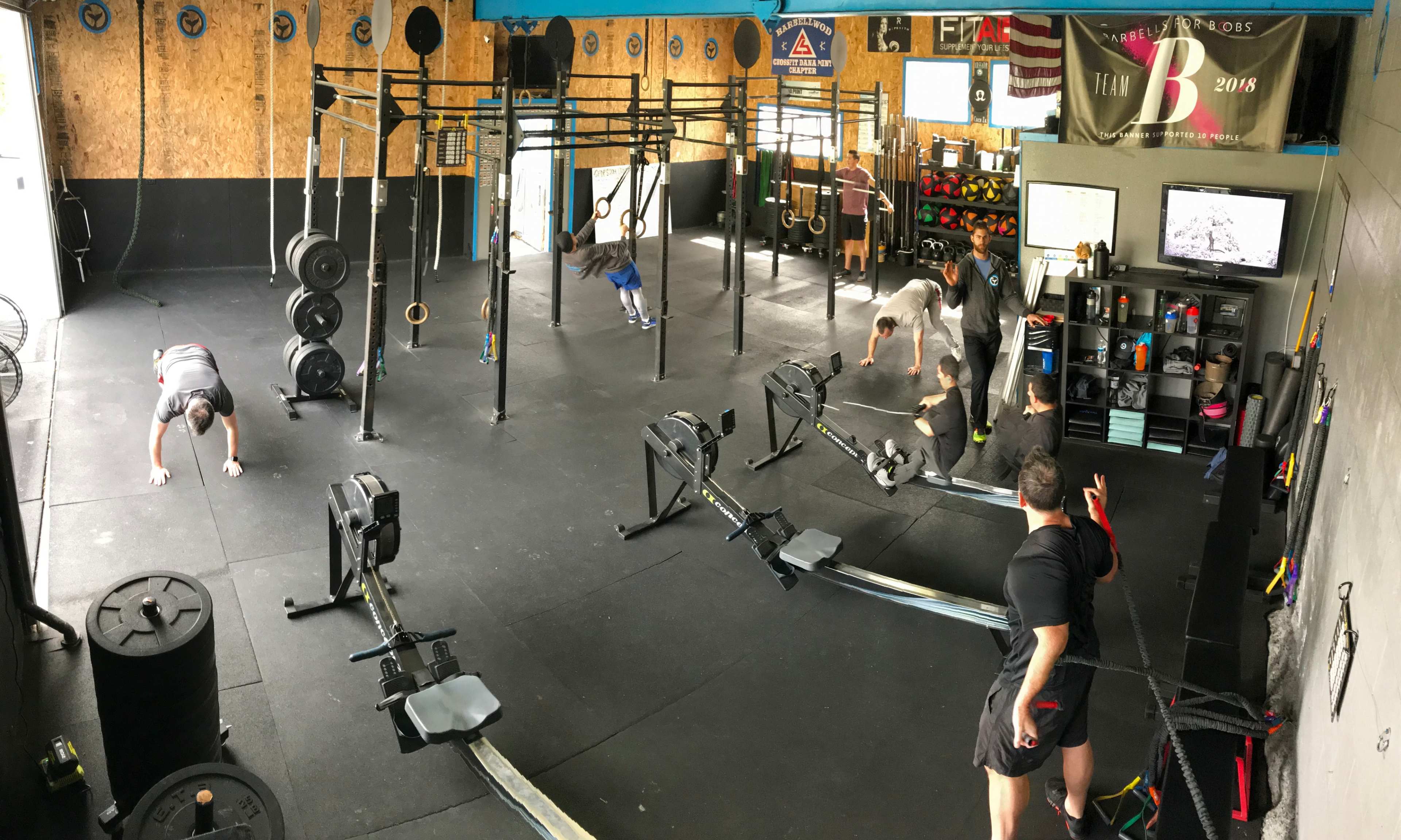 The image shows a gym interior with multiple exercise stations, including rowing machines and weightlifting equipment, where several people are engaged in various workouts.
