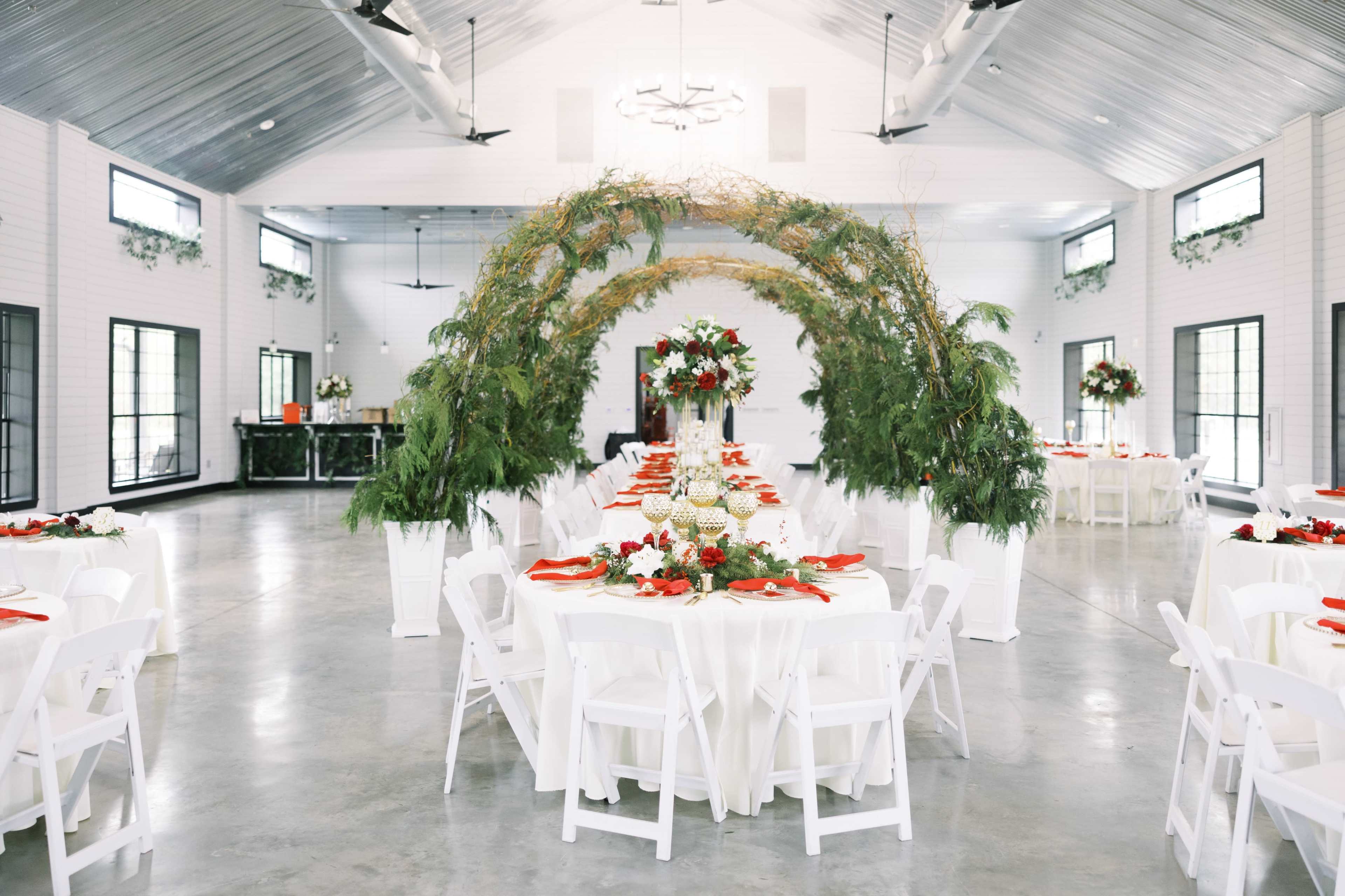 A large event space features elegantly arranged dining tables under a decorative arch made of greenery and flowers.