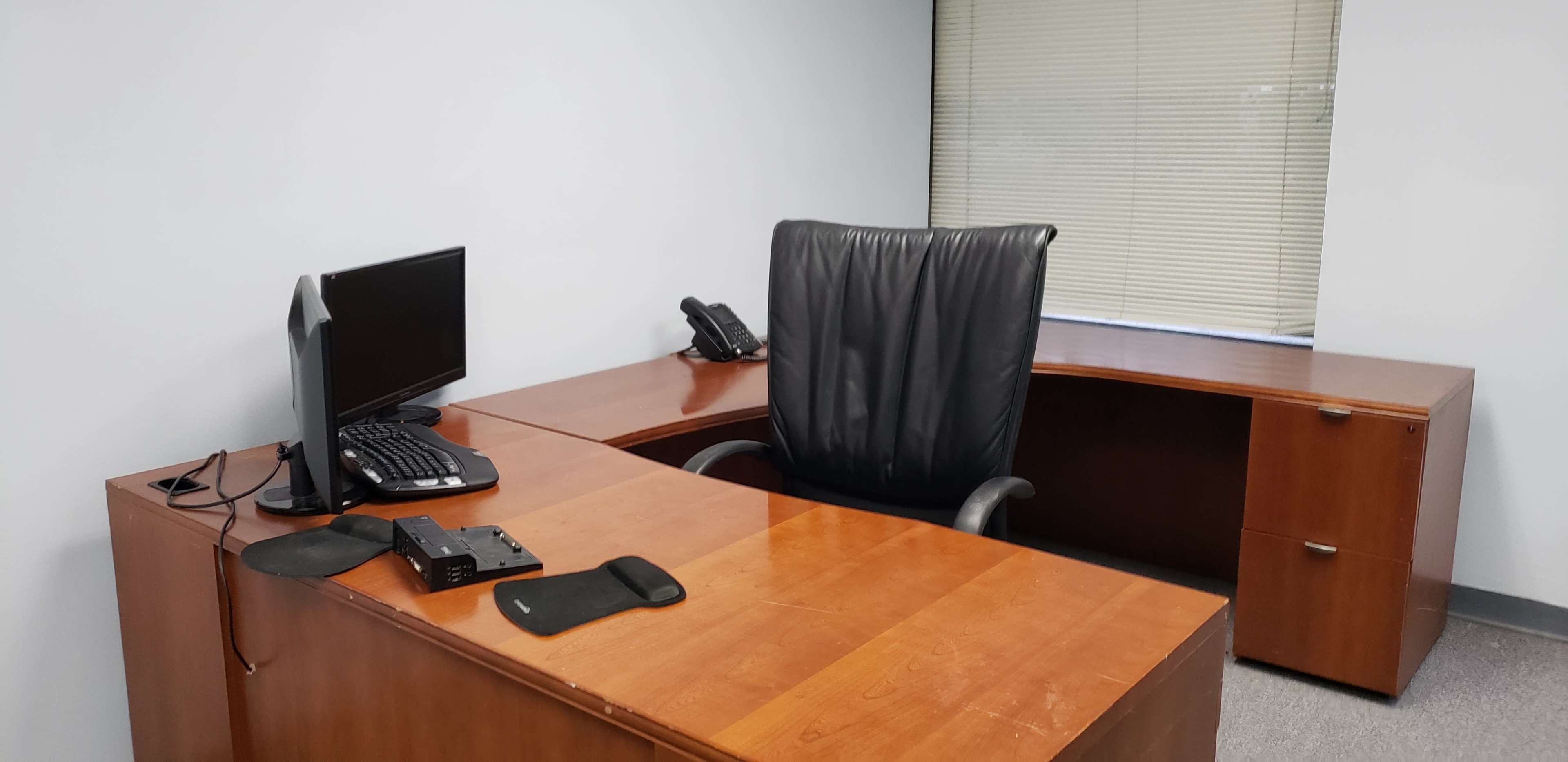 A minimally furnished office with a wooden desk, a black ergonomic chair, a computer monitor, a keyboard, a telephone, and a file cabinet.