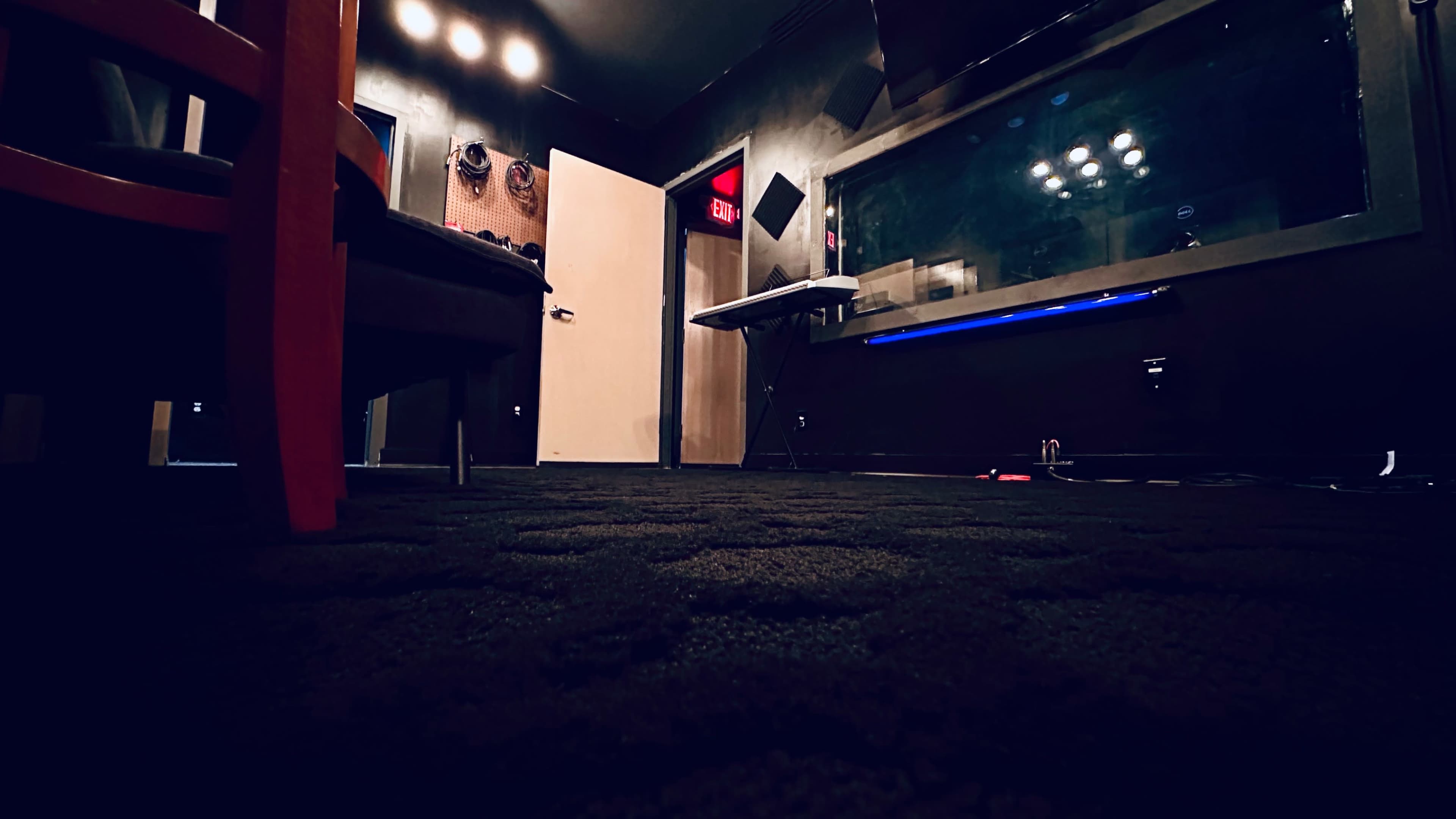 A recording studio with a dark carpeted floor, a door partially open, and a control room visible through a glass wall.