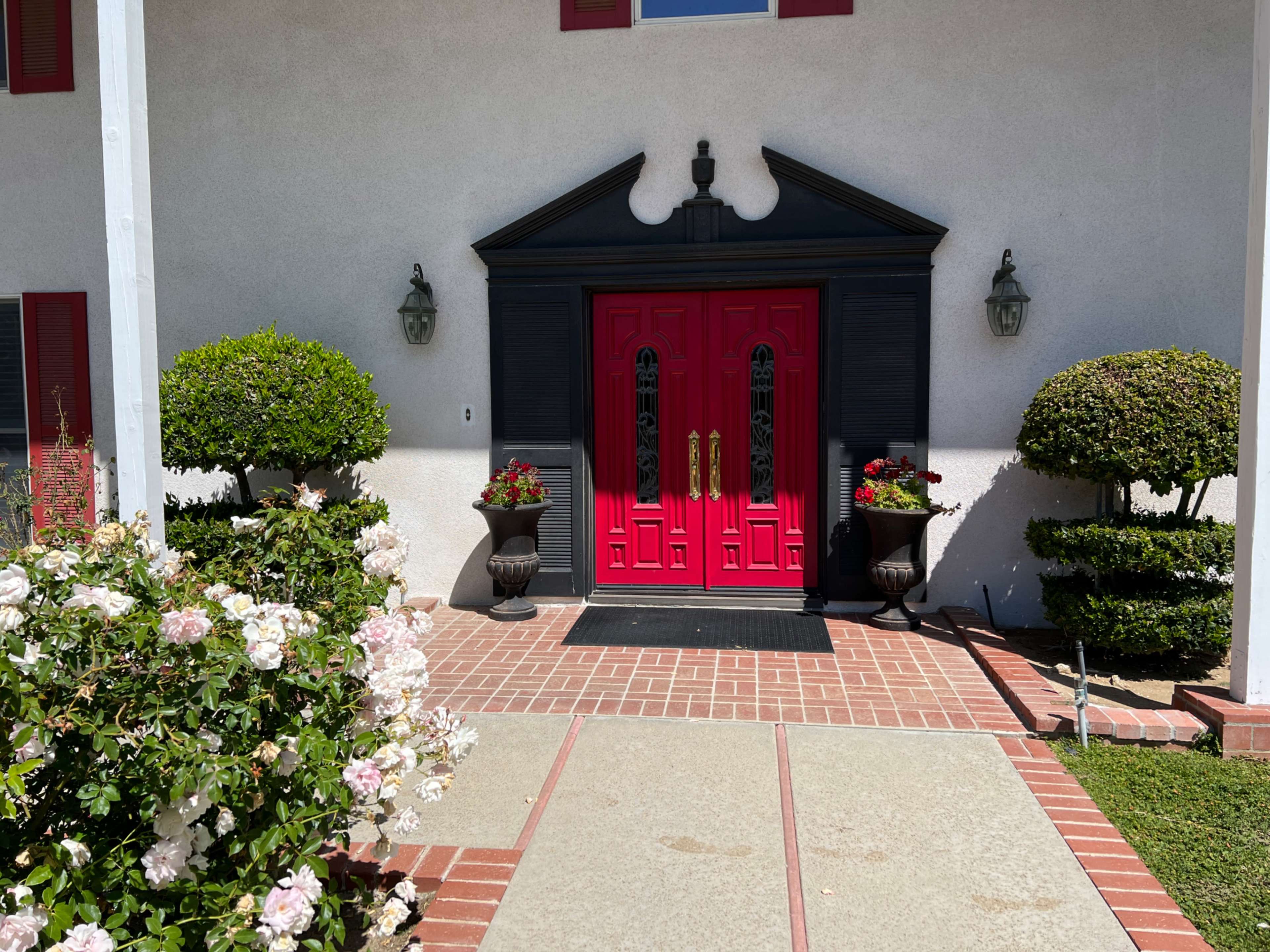 A white house featuring a black-trimmed entrance with a bright red double door, flanked by potted plants and surrounded by manicured hedges and flowers along a stone pathway.