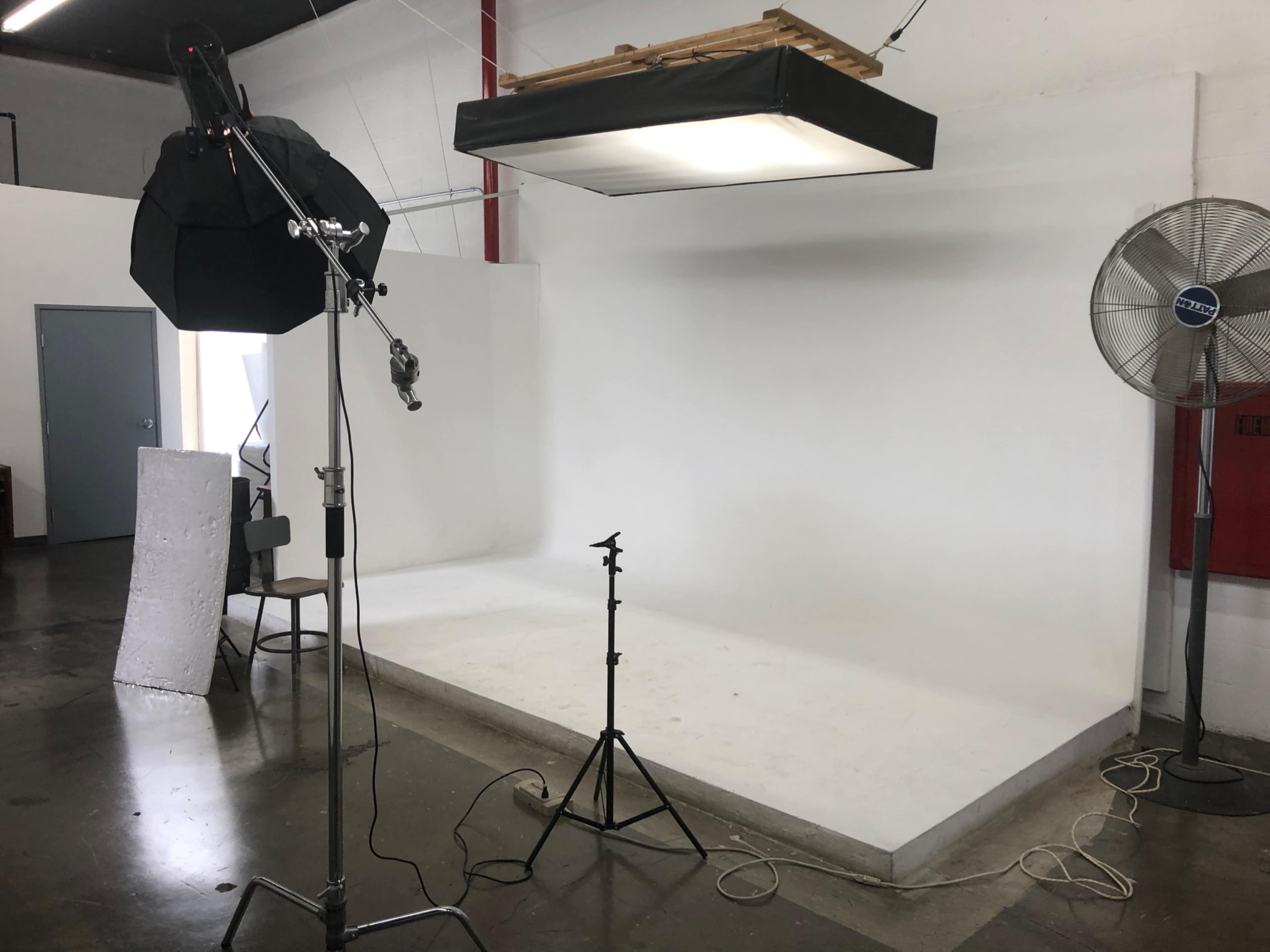 A photography studio with a white backdrop, lighting equipment, a fan, and a chair.