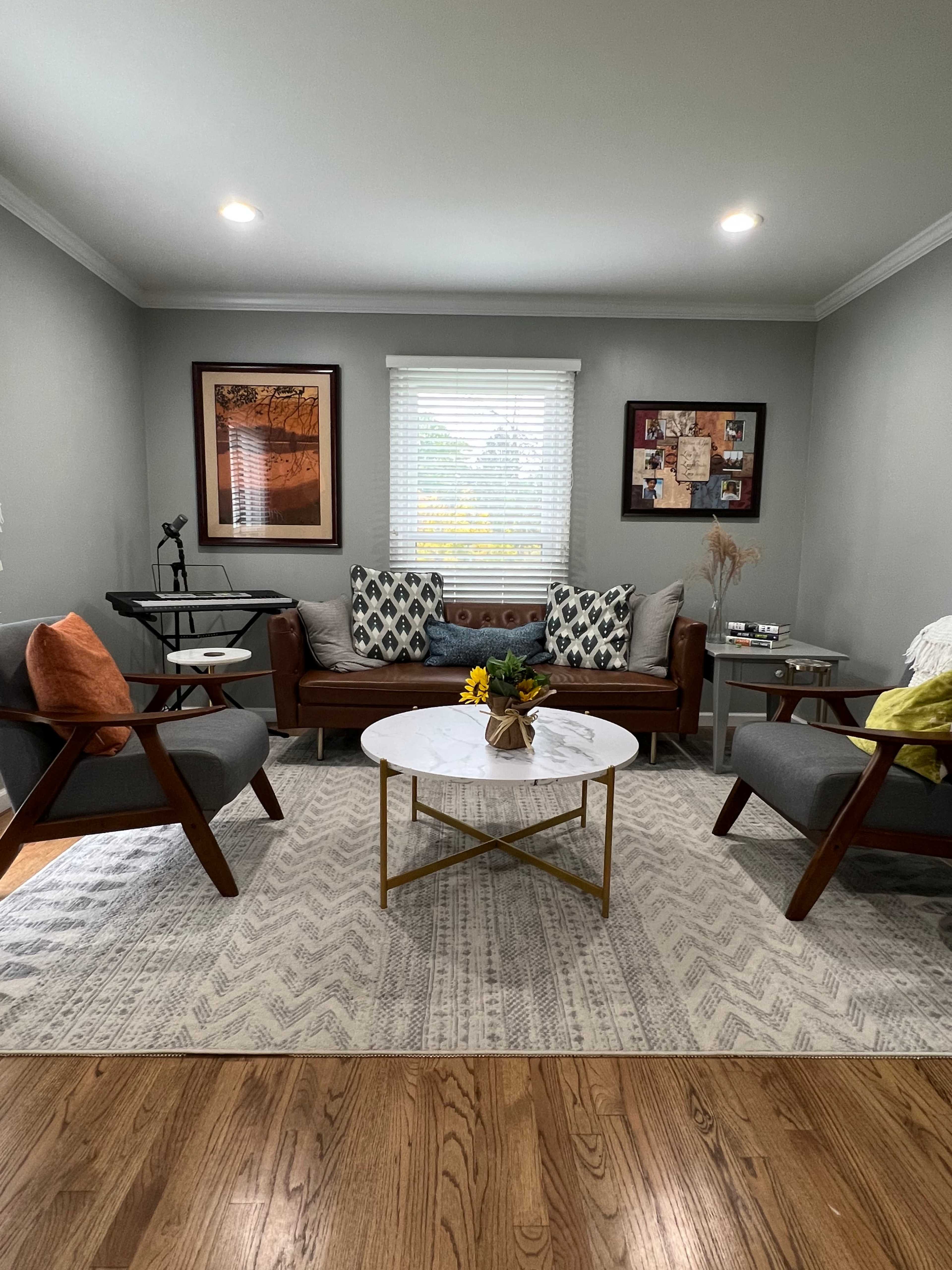 The image shows a living room with a brown leather sofa, two gray armchairs, a round coffee table with a floral arrangement, and framed artwork on the walls.