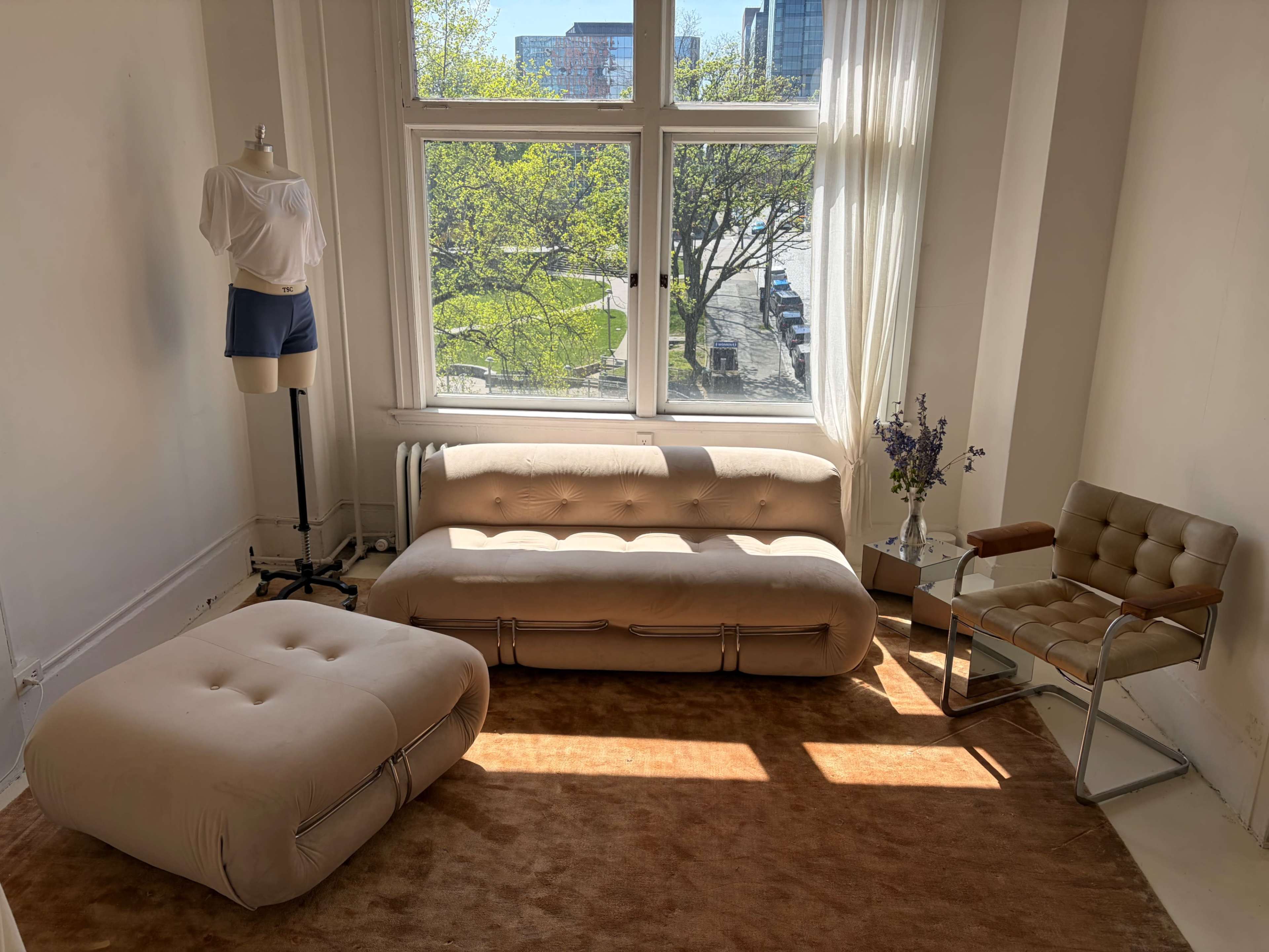 The image shows a well-lit room with a beige upholstered sofa, an accent chair, a plush footstool, a mannequin in minimalist clothing, and a view of a green area outside through large windows.
