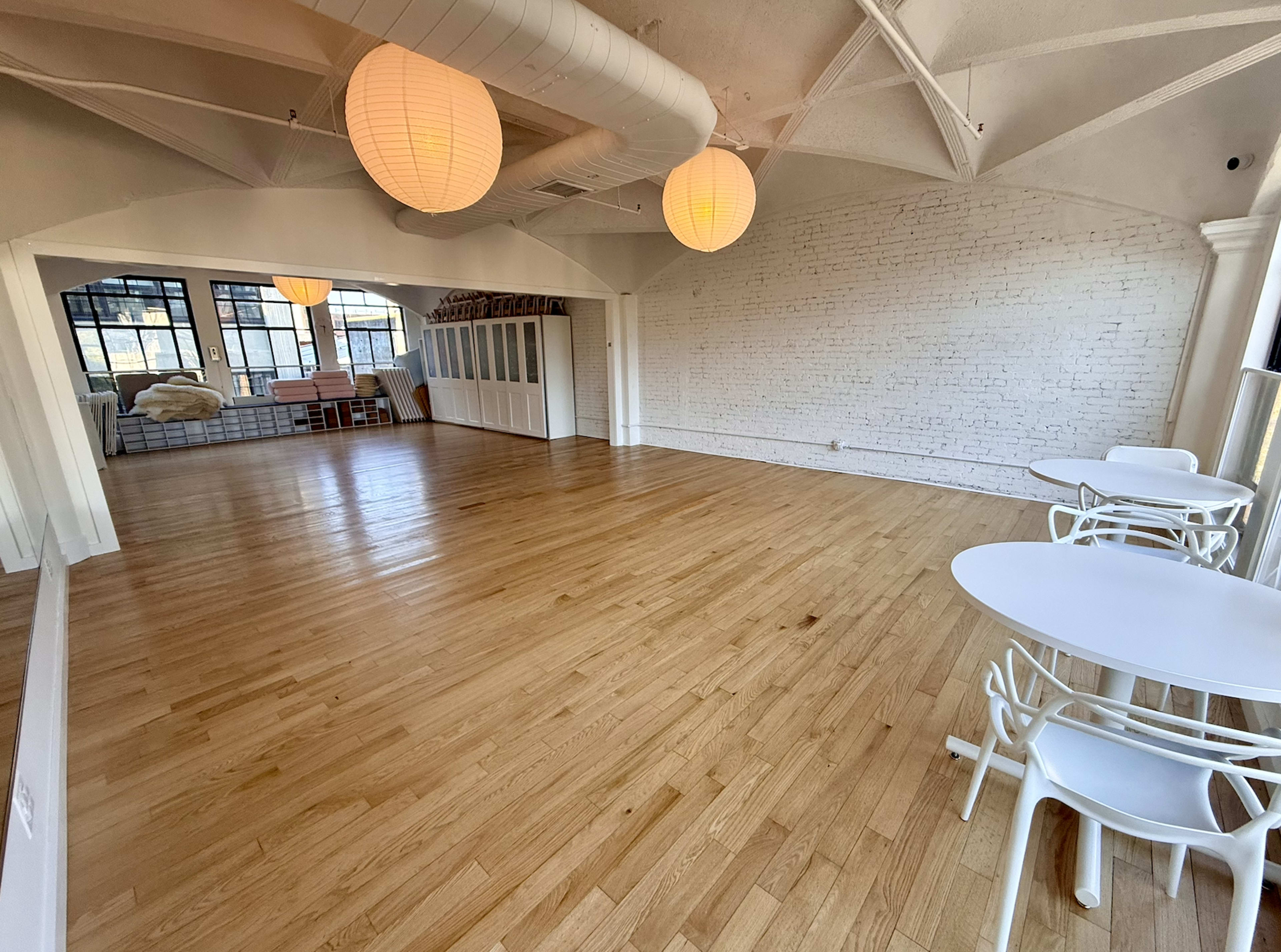 The image shows a spacious, empty room with wooden flooring, two paper lanterns hanging from the ceiling, and white brick walls.