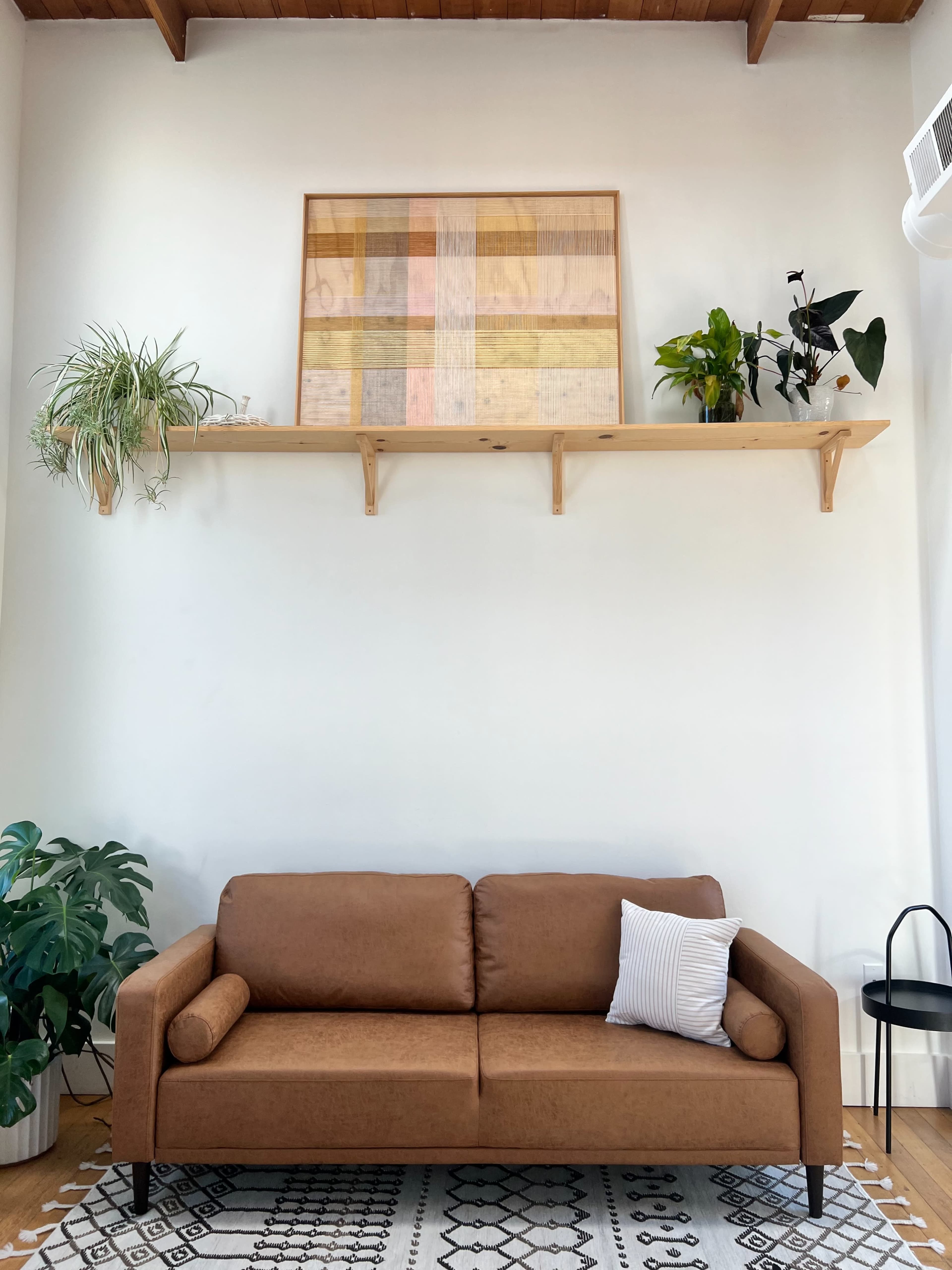 The image shows a brown sofa placed in front of a white wall, with a wooden shelf above it holding plants and artwork.