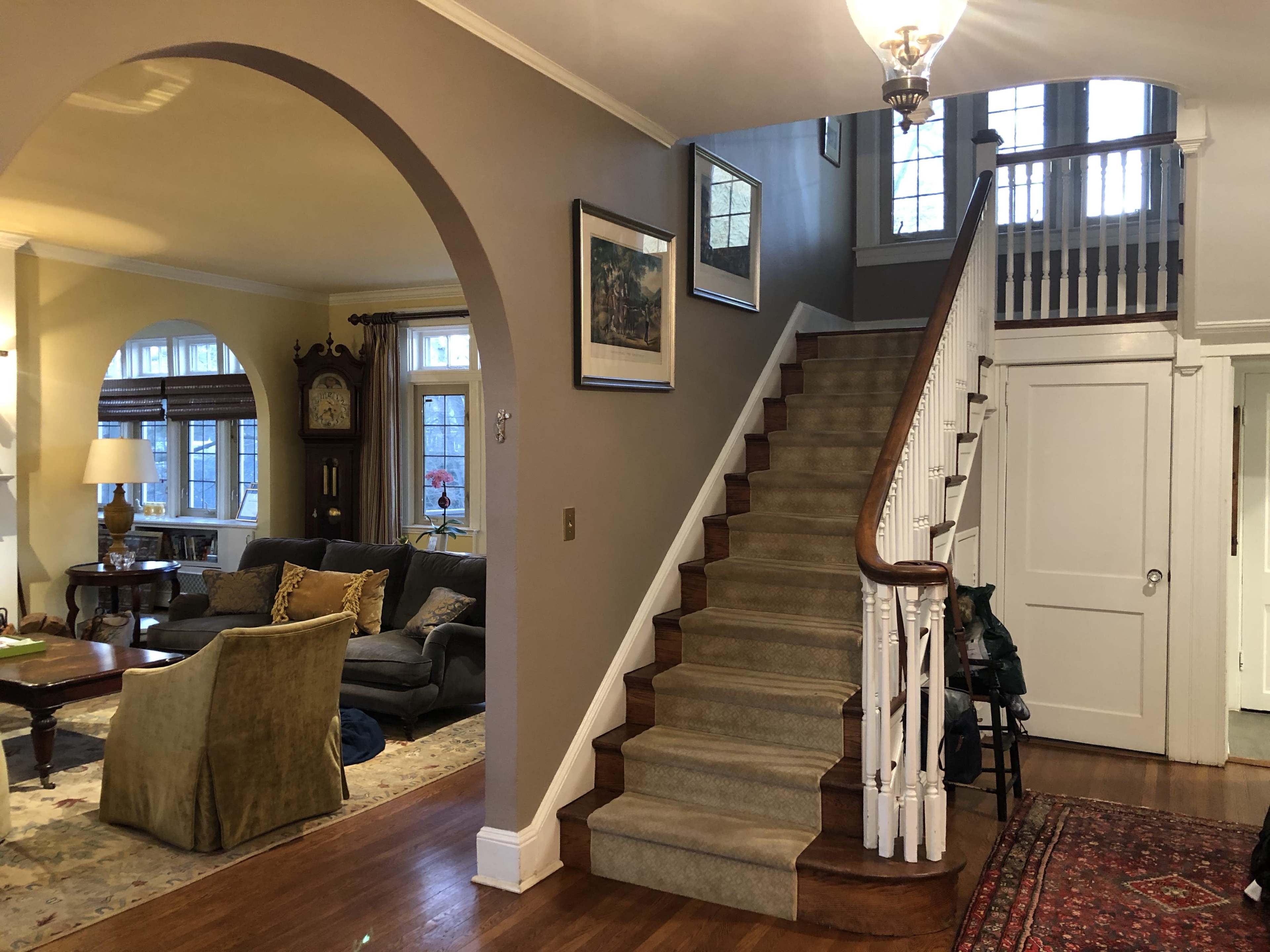 The image shows a domestic interior featuring a staircase leading to an upper level, with a living area visible on the left side and a foyer with a chandelier.