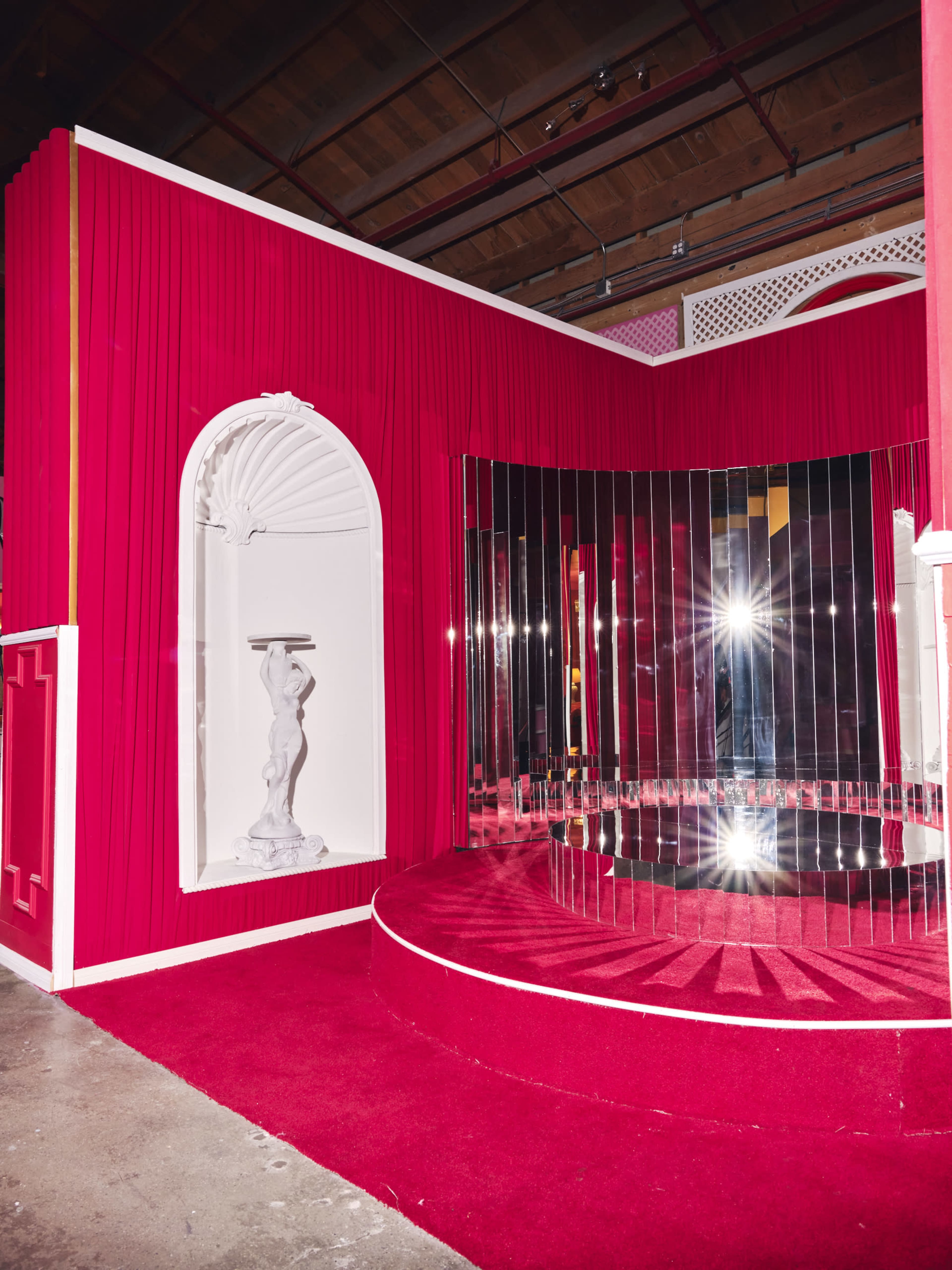 A vibrant pink room featuring a mirrored backdrop, a circular platform, and a white sculpture set in an archway.
