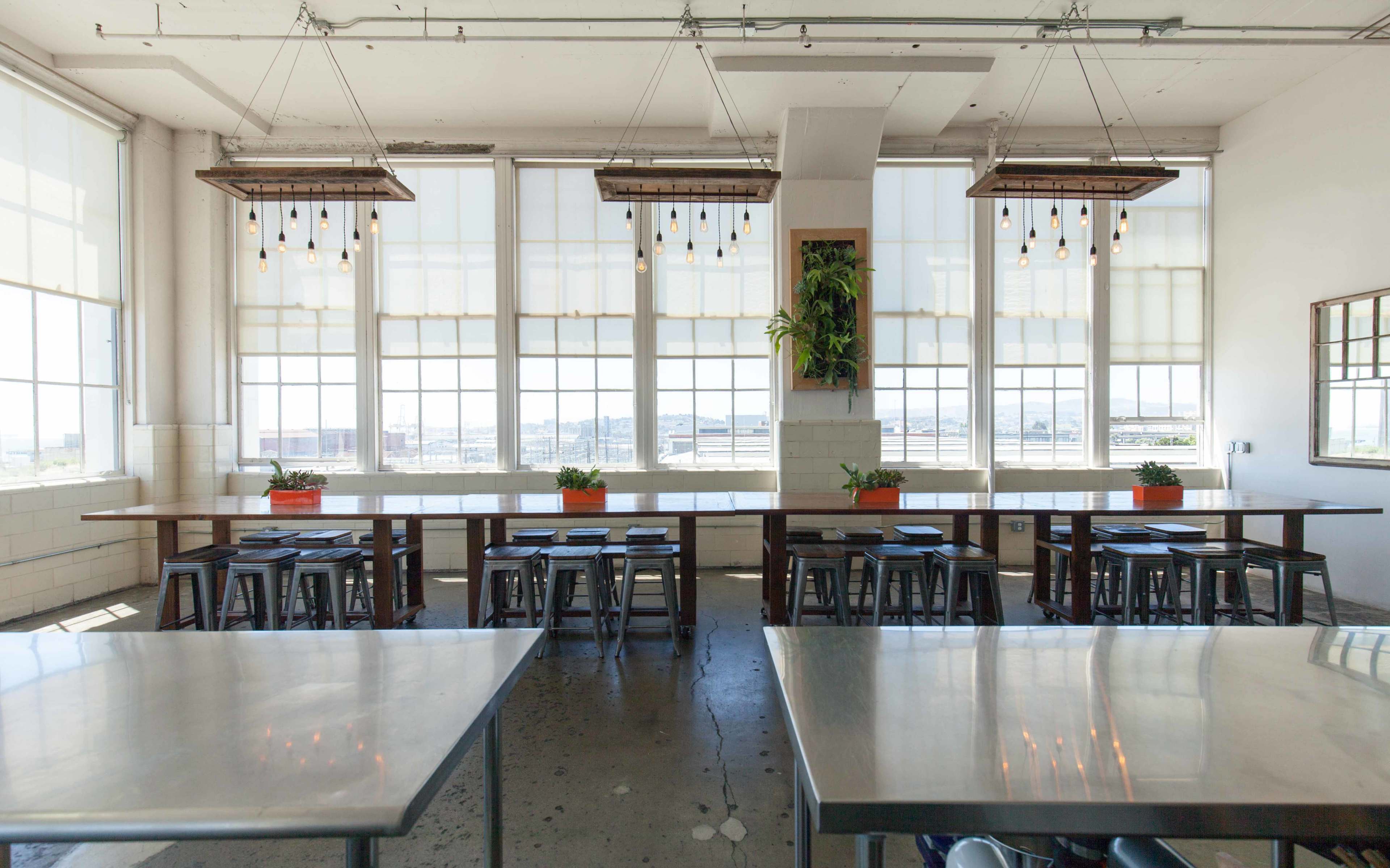 A spacious dining area with long wooden tables, metal stools, and large windows that offer a view of the outdoors, decorated with hanging light fixtures and potted plants.