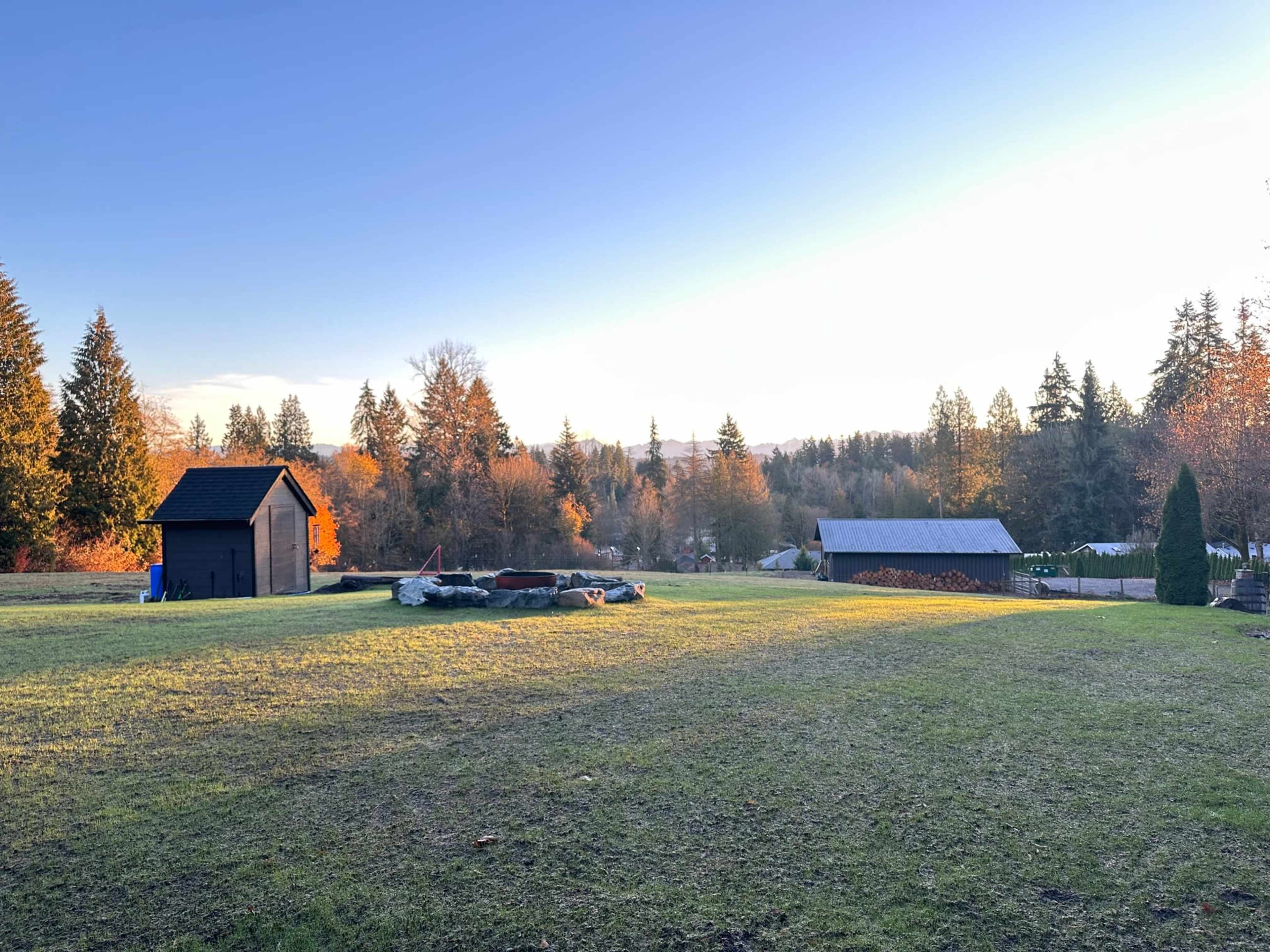 The image shows a grassy field at sunset, with a small black shed and a larger gray building in the background among trees with autumn foliage.