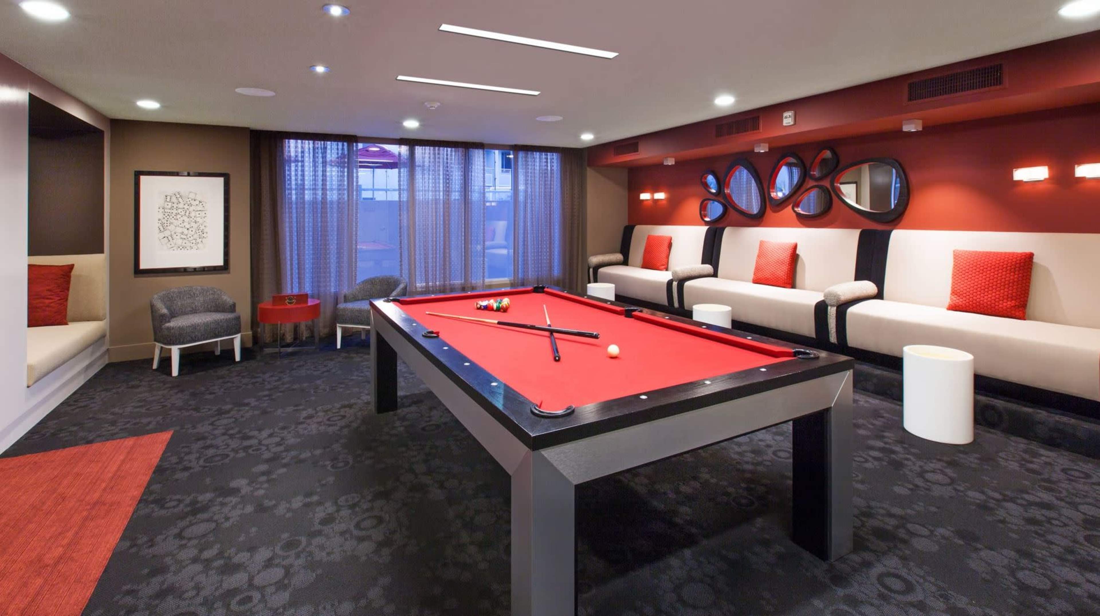 The image shows a game room featuring a red pool table, surrounded by seating areas with modern decor and ambient lighting.