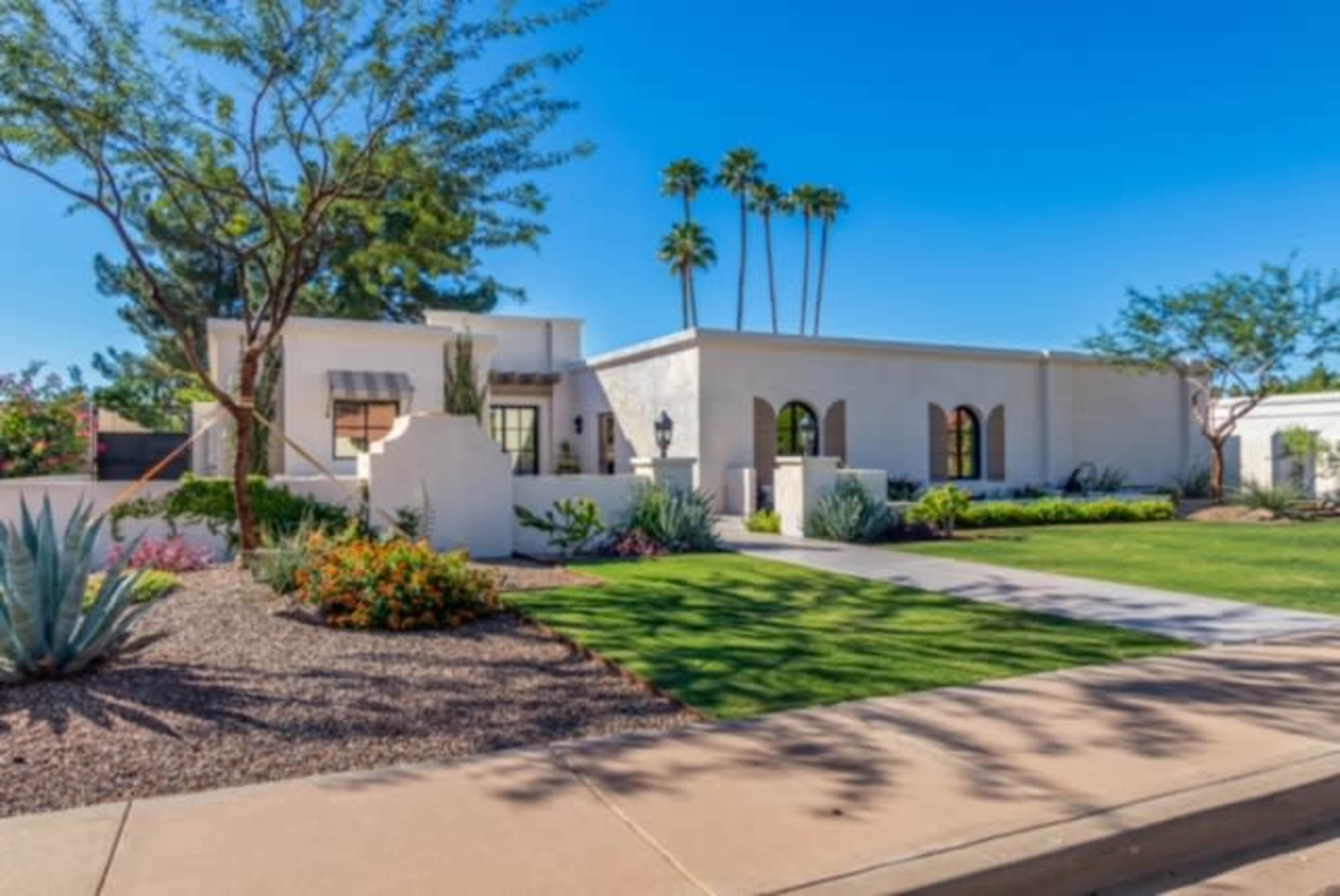 A single-story white stucco house with large windows is surrounded by manicured lawns and desert landscaping, featuring palm trees and colorful shrubs.
