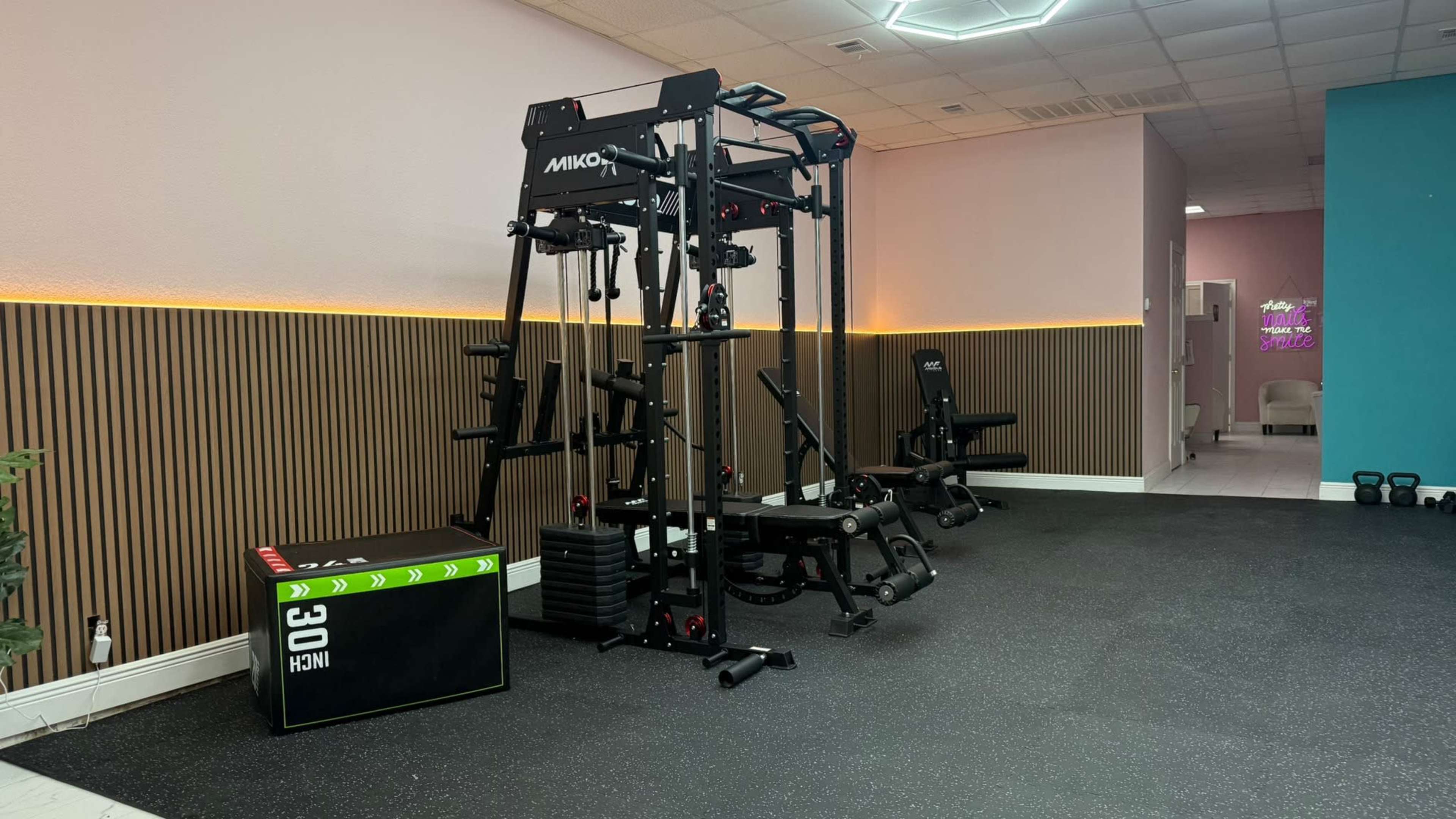 The image shows a modern gym interior featuring a multi-functional exercise machine, a weight bench, and a set of kettlebells on a rubber floor.