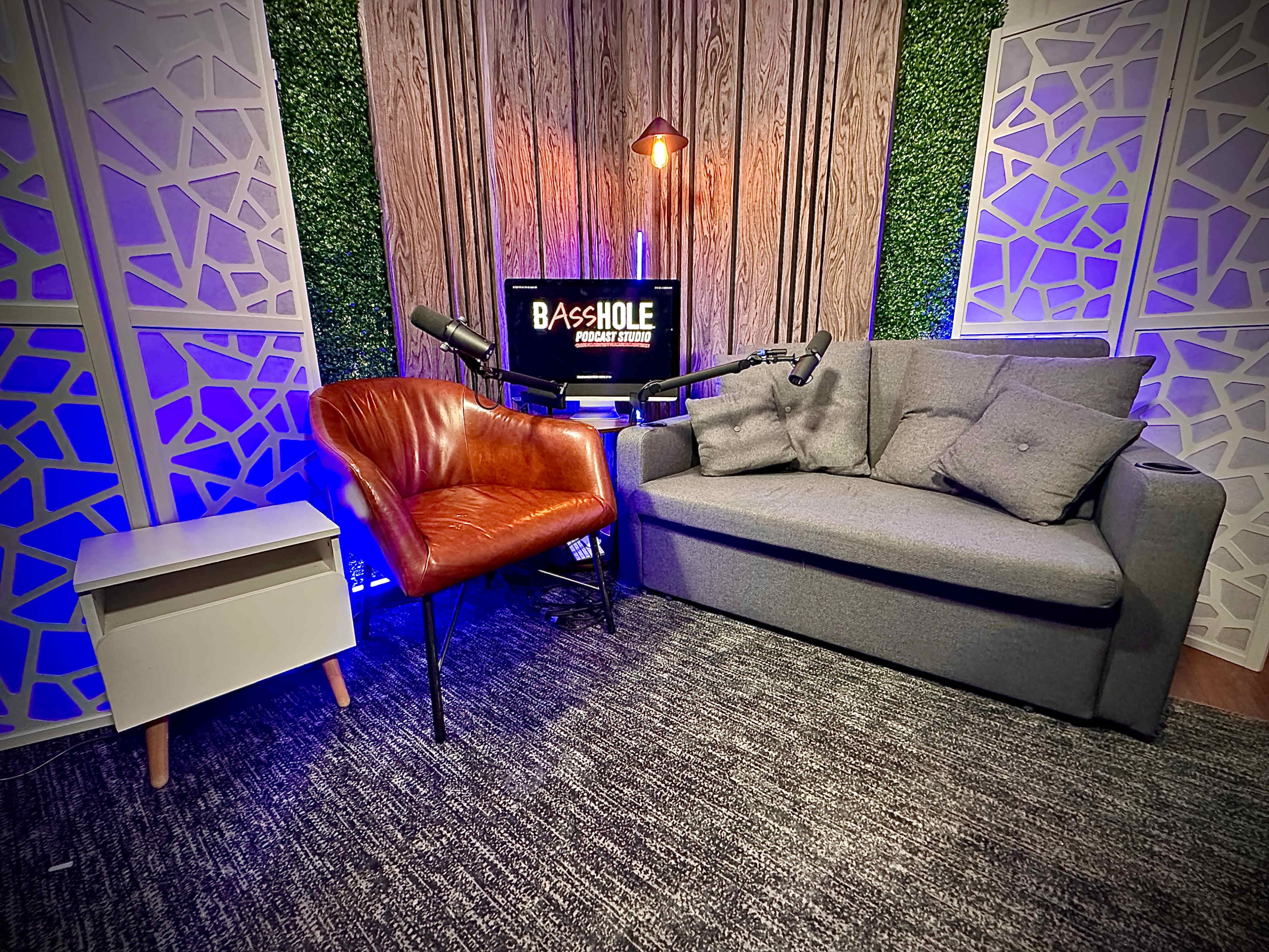 The image shows a podcast recording setup featuring a gray sofa, a brown accent chair, a small white side table, and a screen displaying the text "BASSHOLE Podcast Studio" against a backdrop of decorative wood and greenery.