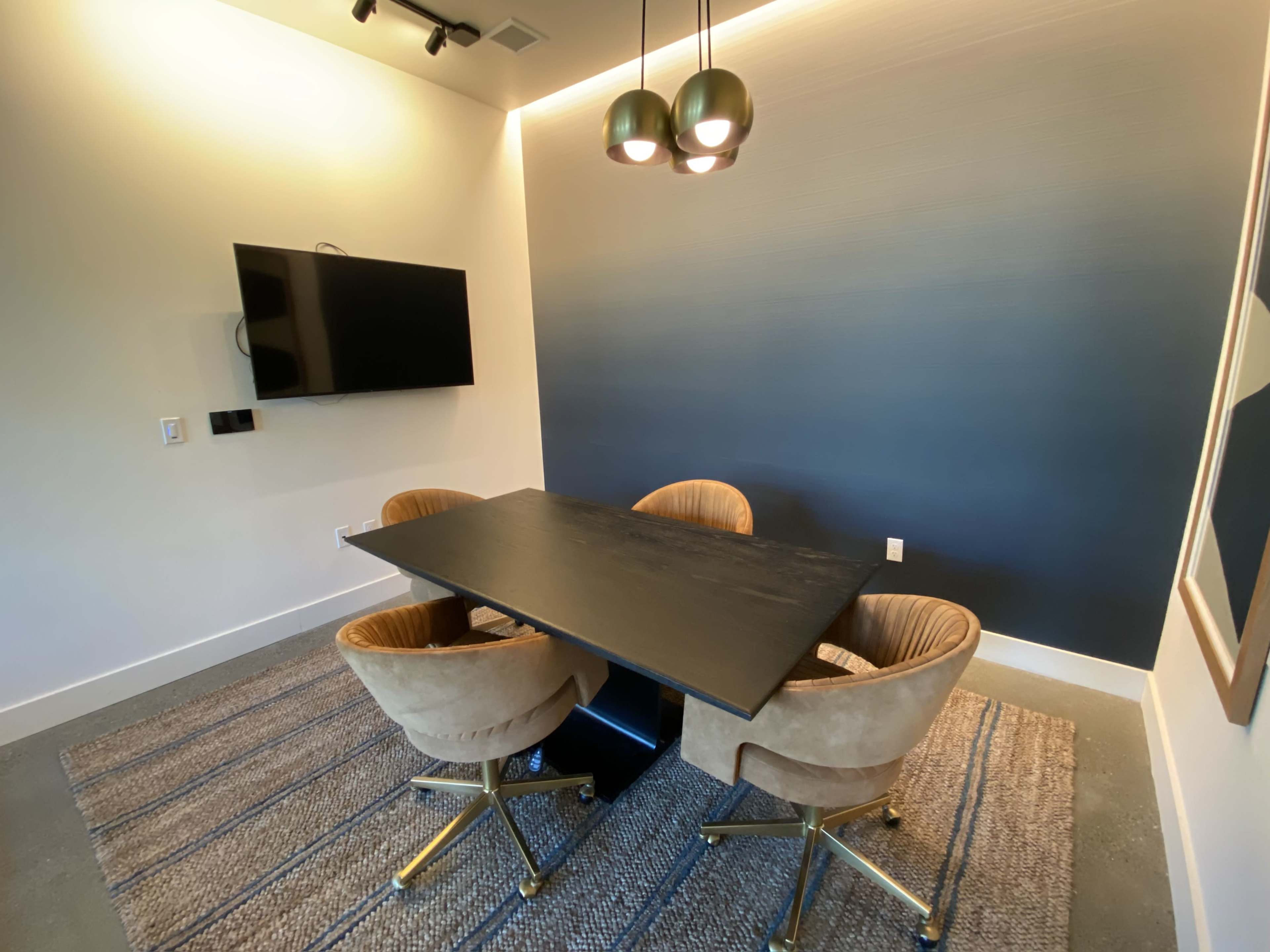 A modern meeting room features a dark table with four round chairs, a wall-mounted television, and a gradient blue wall.