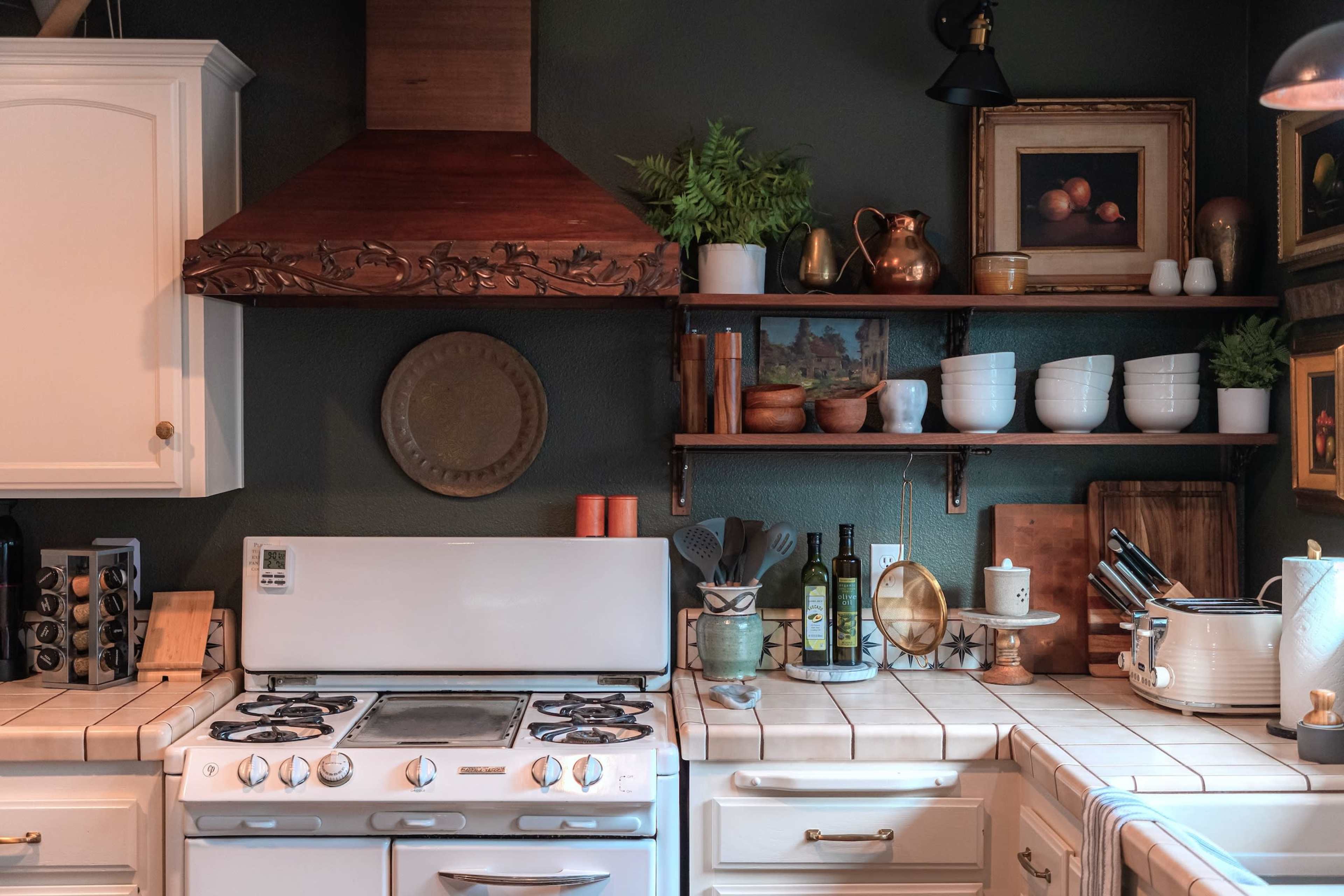 A cozy kitchen features a white stove, decorative shelves with plants and dishware, and a wooden vent hood above the countertops.