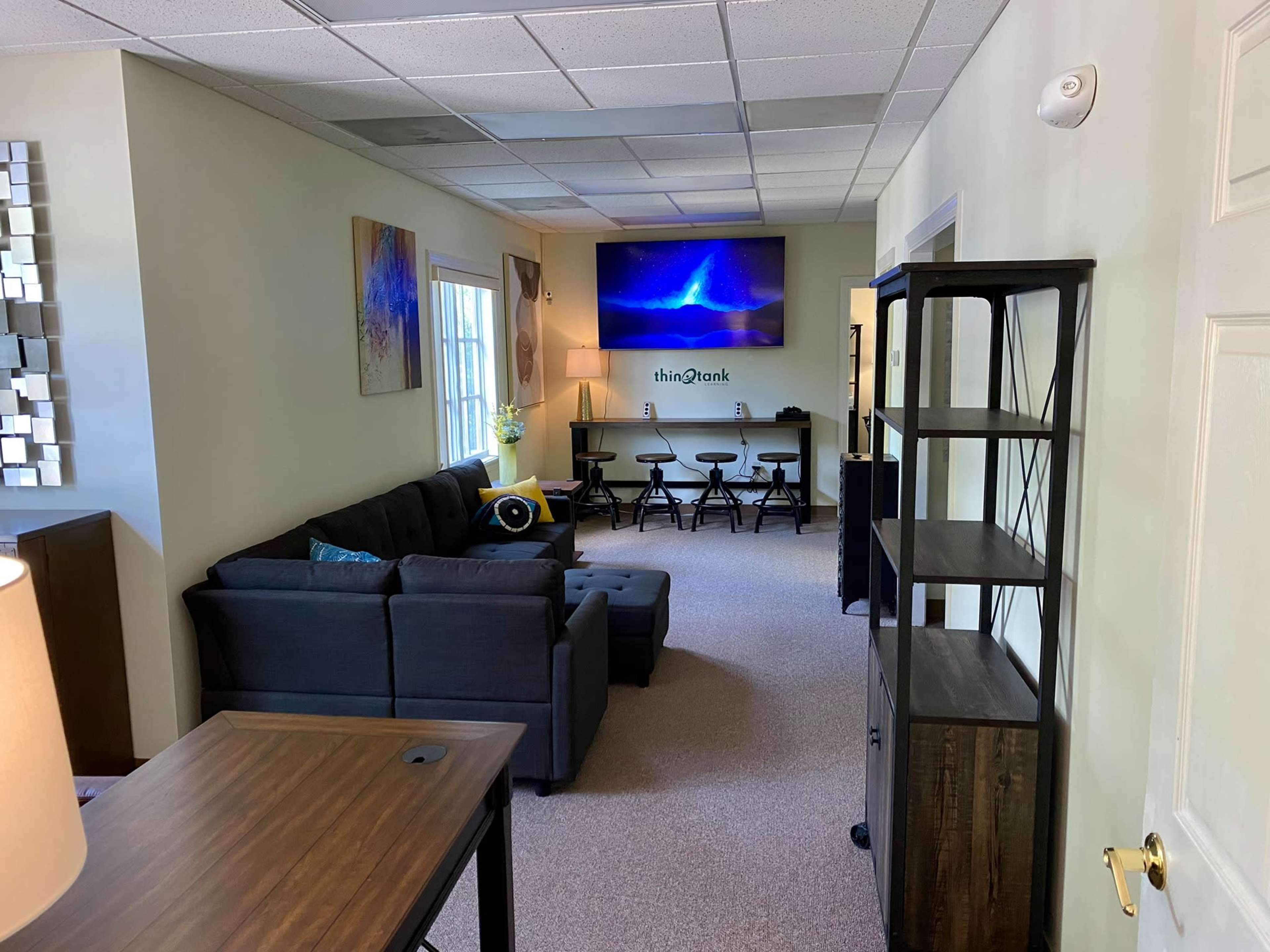 A modern office space with a sectional couch, a large television mounted on the wall, a small table with stools, and a bookshelf.